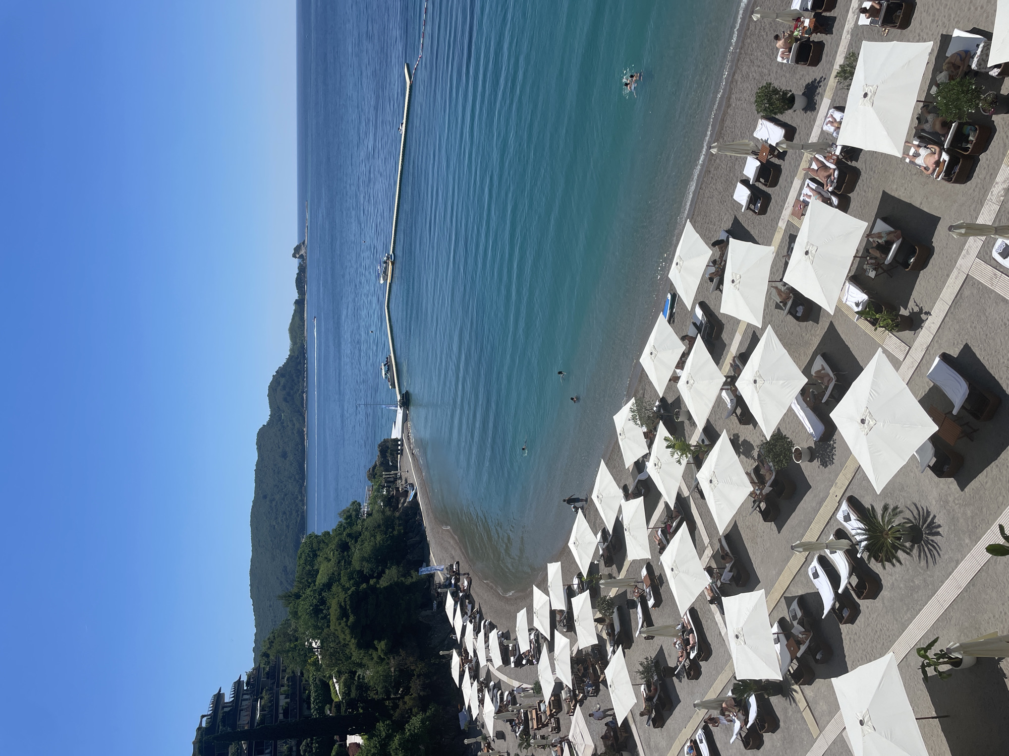 This aerial shot captures a beach in Budva, Montenegro. The scene includes a sandy beach populated with rows of white umbrellas and lounge chairs where people relax. The Adriatic Sea is visible, with swimmers and a long floating pier extending into the water. In the background, a hillside covered with trees and buildings rises from the coast. The sky is clear and blue.