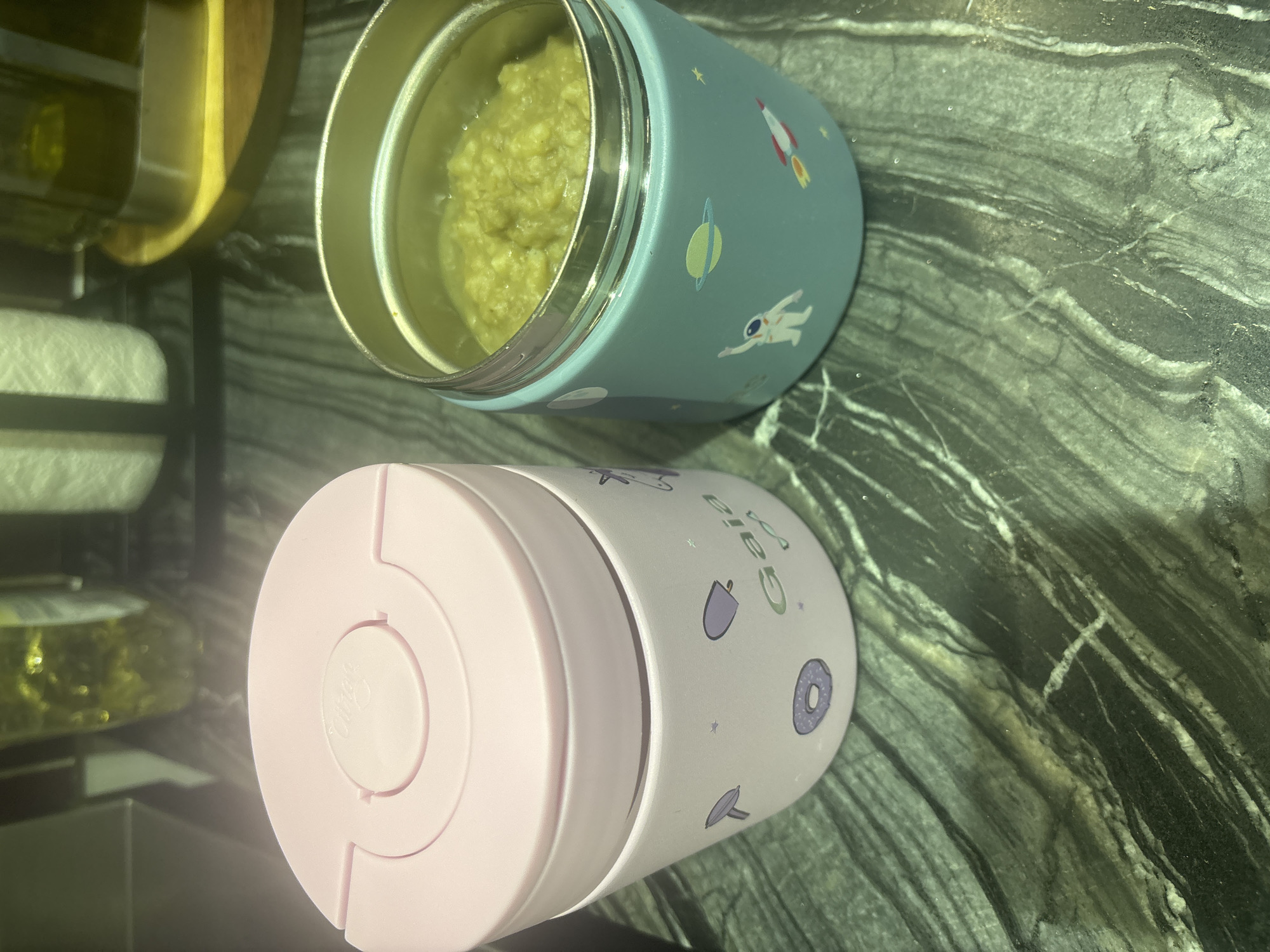 Two insulated food jars sit on a dark green, veined countertop. The jar on the left is pink with a lighter pink lid. Its side is white and decorated with purple illustrations of a lollipop, a donut, a unicorn, and stars, along with the word "Gaia". The jar on the right is teal and filled with a textured, light green substance. It is decorated with illustrations of an astronaut, a planet, and a rocket. The background shows blurred objects, including bottles and what appears to be a roll of paper towels, suggesting an indoor setting, likely a kitchen or pantry. No specific landmarks or contextual clues indicate the city of Dubai, United Arab Emirates.