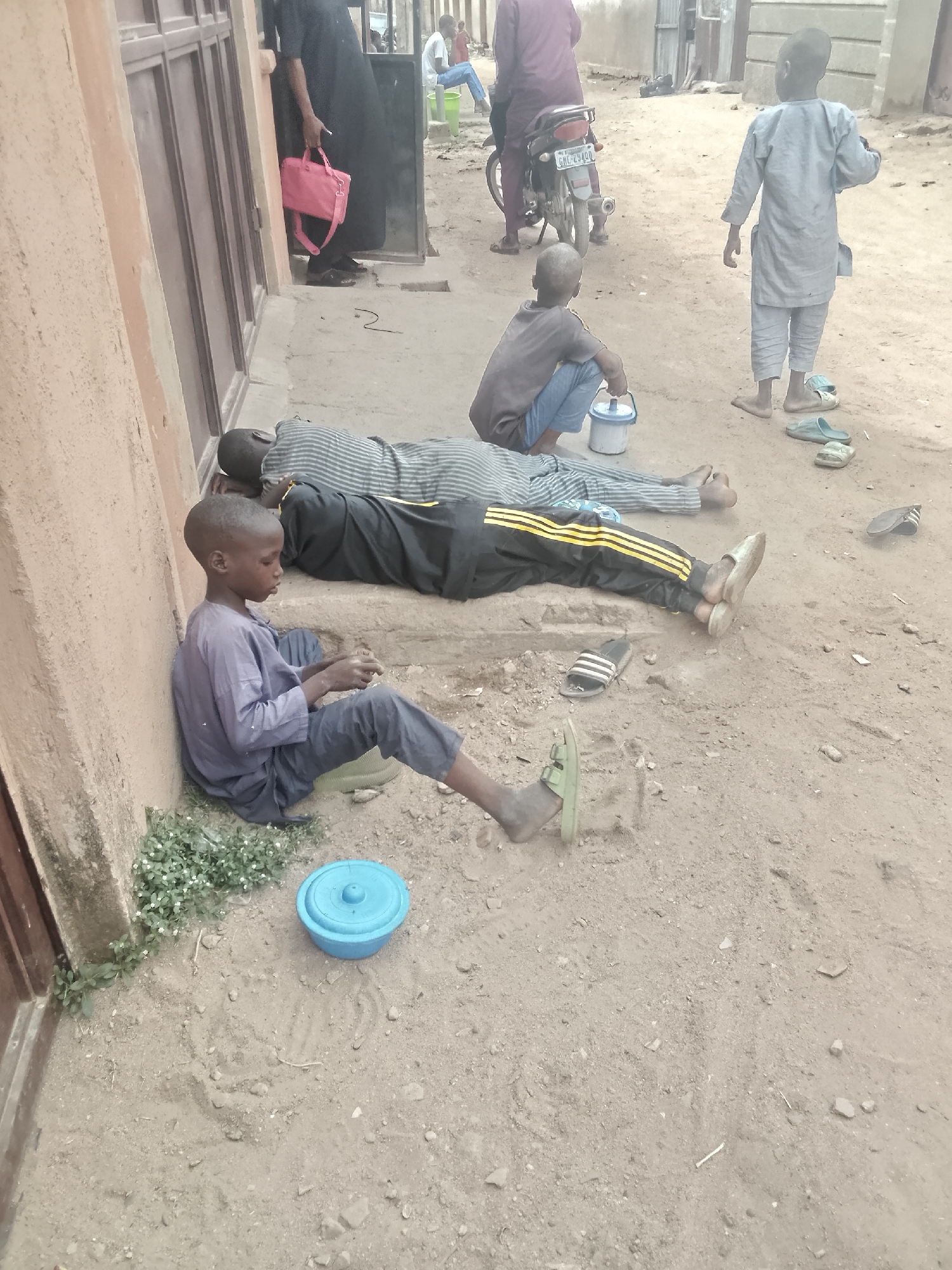A street in Pantame, Nigeria, features multiple people. A boy is sitting with his back against a wall, and a blue container is near him. Next to him, two people are lying down. Nearby, another boy is sitting next to a white container. Further away, a boy is standing and wearing sandals. In the background, there is a parked motorcycle and two people in the distance. One person is carrying a pink bag. The ground is sandy and uneven.