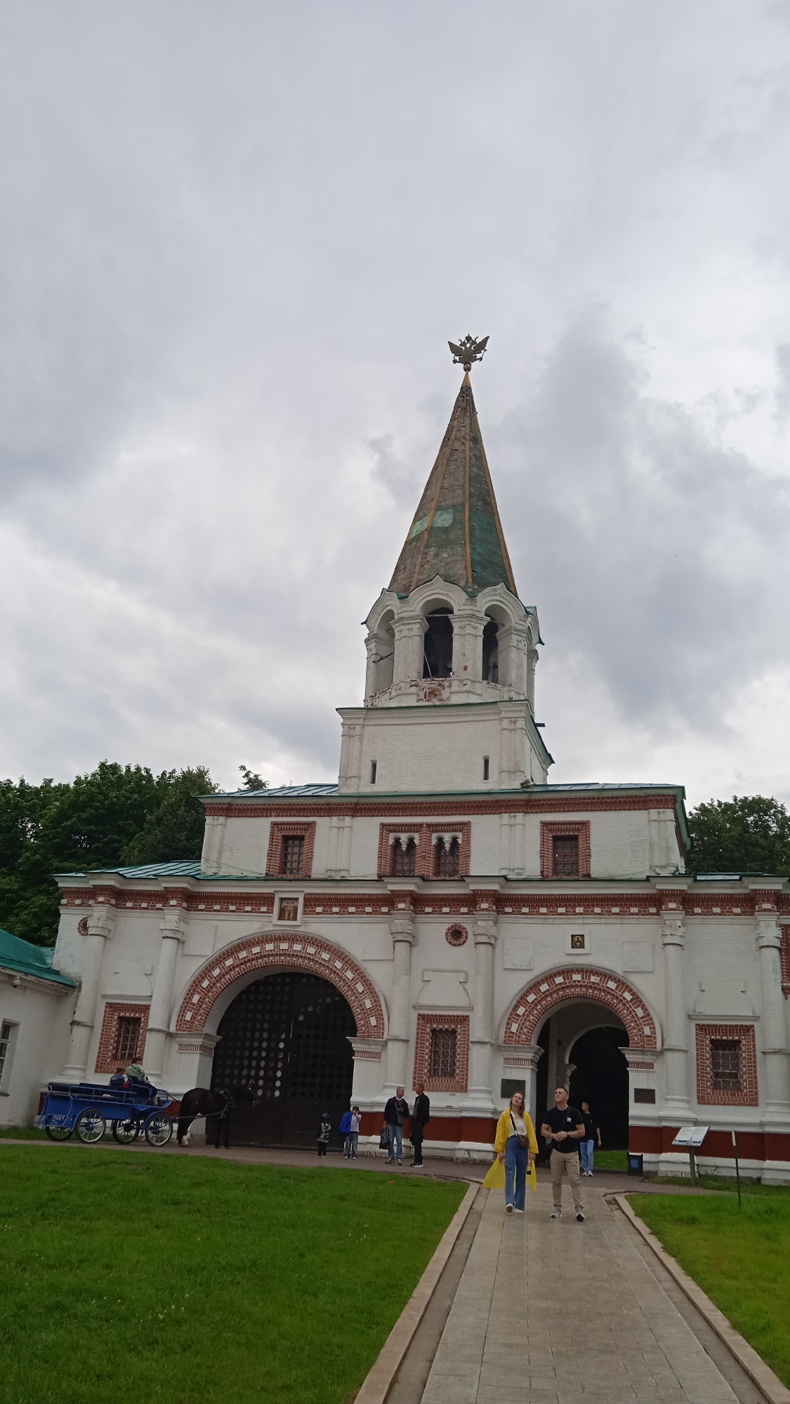 The image depicts an outdoor scene of a grand, historic architectural structure, possibly a gatehouse or bell tower, under a cloudy sky. The building is predominantly white with red brick accents and decorative elements. A tall, slender spire with a double-headed eagle crest crowns the structure. Arched doorways and windows are visible, along with intricate brickwork patterns.
In the foreground, a paved pathway leads towards the building. On the left side of the path, a horse-drawn carriage, painted blue, is parked on a grassy area. Several people are present in the scene. Two individuals, a woman in a yellow jacket and jeans and a man in a dark t-shirt and light pants, are walking along the path. Other individuals are visible near the large arched entrance, including children and adults. One person appears to be operating the horse-drawn carriage.
The weather appears overcast, indicated by the gray, textured clouds covering the sky. The greenery of trees and grass suggests a temperate climate, and the overall atmosphere is that of a tourist or public space, likely a historical site. No visible text is discernible in the image. The time of day appears to be daytime, given the ambient light. The people's expressions are not clearly discernible to infer specific emotions.