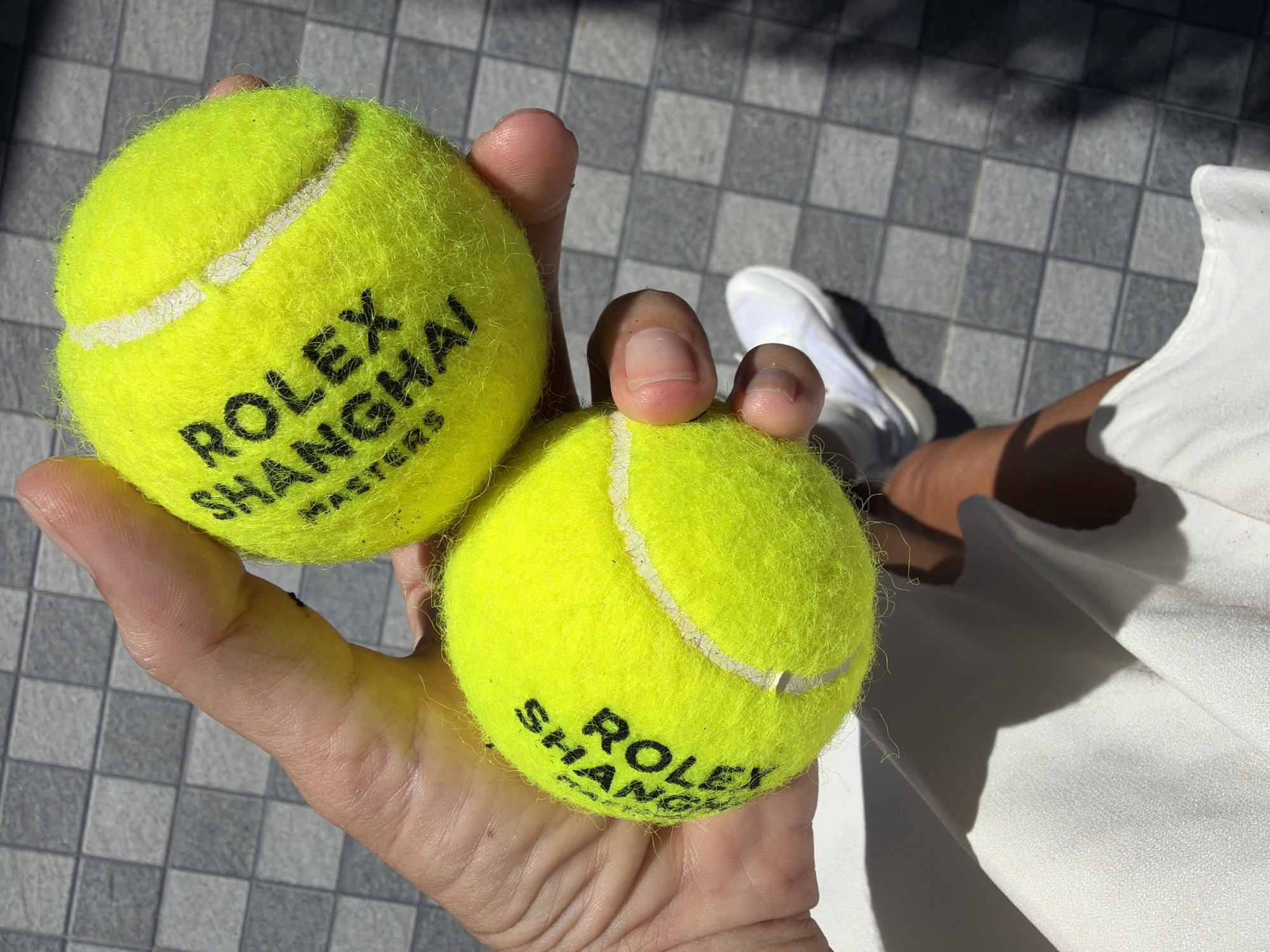 The image shows a person holding two bright yellow tennis balls, each with black text that reads "ROLEX SHANGHAI MASTERS". The balls are fuzzy, typical of tennis equipment. The person's hand, with visible fingers and fingernails, is holding the balls, suggesting they are the subject's primary focus. In the background, the ground appears to be tiled with a gray and white checkered pattern, consistent with an outdoor setting. A white athletic shoe is also visible in the background, implying the person is wearing athletic attire.
The lighting suggests it is daytime, with harsh shadows cast on the ground, indicating bright sunlight. The overall scene is casual and appears to be set in a location where tennis is played or has recently been played. The presence of the "Rolex Shanghai Masters" text indicates a connection to a professional tennis tournament. While the exact activity isn't explicitly shown, the context implies participation in or association with the tournament. The mood seems relaxed and perhaps celebratory, given the close-up view of the branded items. The location context of Baan Bo Phut, Thailand, is noted but not visually evident in the provided image itself.