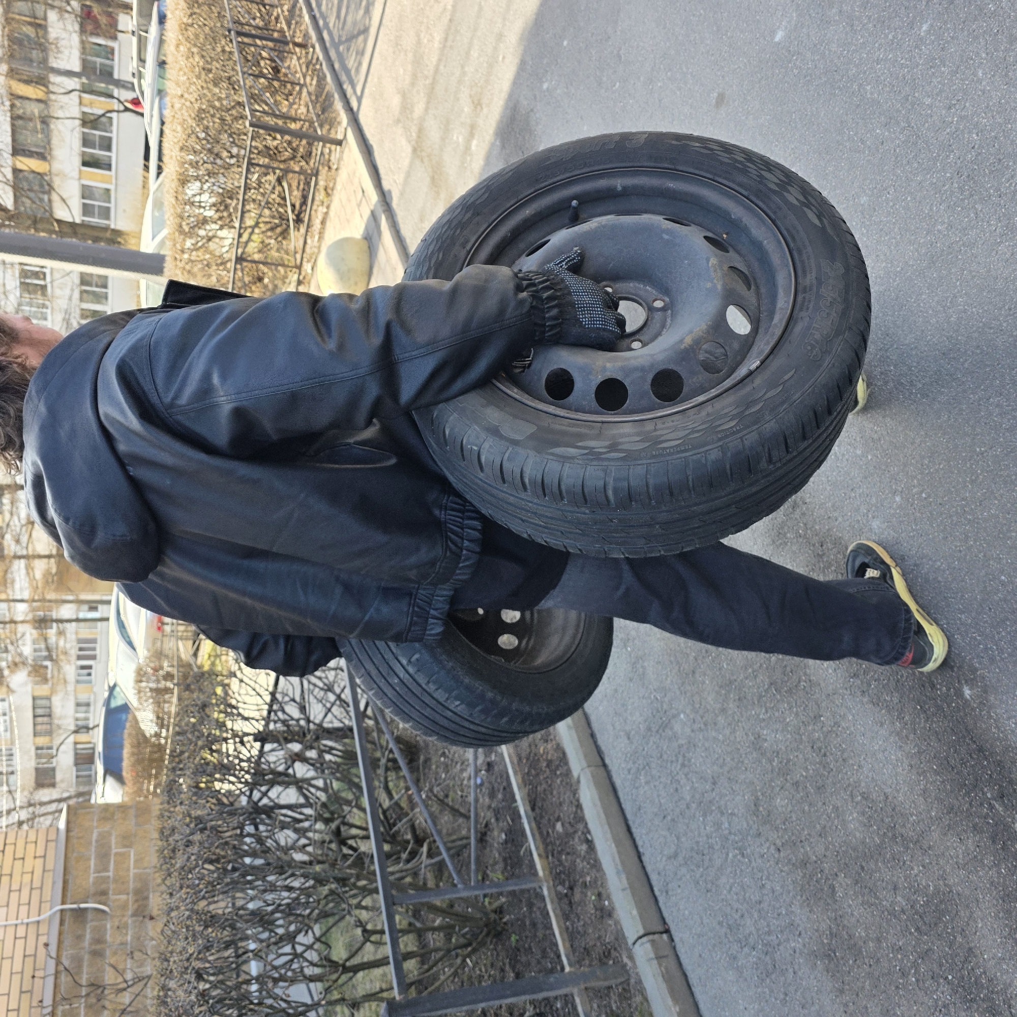 A person is carrying two tires on their shoulder on a paved street. The person is wearing a black leather jacket, black jeans, and black sneakers with a yellow stripe. They are also wearing gloves. The tires are black and have steel rims.
In the background, there is a brick building with a hedge, a metal fence, and several multi-story buildings. The setting appears to be a residential area. Considering the architecture and surrounding environment, it is likely the location is in Saint Petersburg, Russia.
