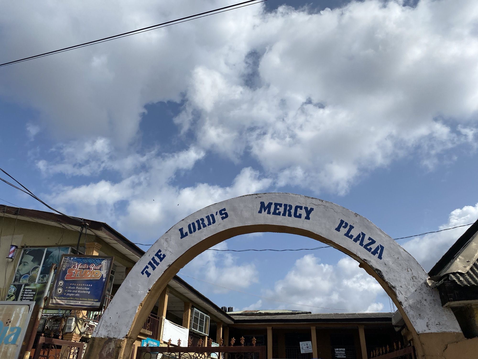 The image shows an outdoor view in Lagos, Nigeria, featuring an arched entrance with the inscription "THE LORD'S MERCY PLAZA" in blue letters. The arch appears weathered with chipped white paint. Adjacent to the arch, on the left, is a building displaying a barber shop sign that reads "Master Beat STUDIO", detailing services like "Barbing," "Hair coloring," and "Dreadlocks," alongside music production services. The contact information indicates the location as Shop & Lord Mercy Plaza, Oja Antop, Epboda, Lagos. The background includes a partly cloudy sky and a building structure with an air conditioning unit.