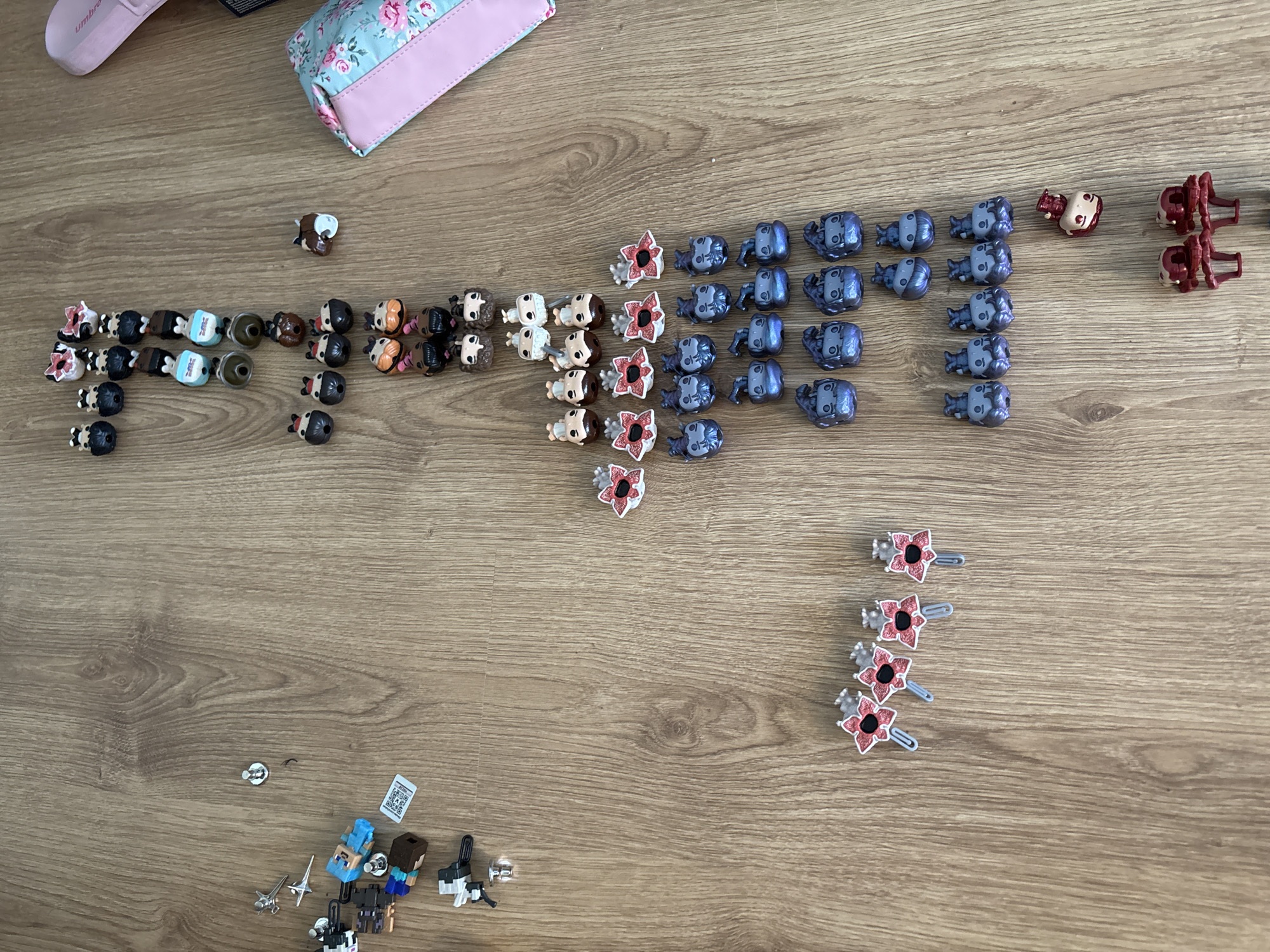 The image shows a collection of miniature figurines and accessories arranged on a wooden surface. The items appear to be organized into rows or groups based on their type or design. At the top left is a pale pink slipper and a small floral patterned purse. A group of Stranger Things figurines including Dustin, Eleven and Will are on the left. Demogorgons are located to the right of the figures, with a single Demogorgon hairclip above 4 others.
To the right are blue figures of an unknown type, and to the right of them, red figures.
At the bottom left, partially obscured, is a collection of Minecraft characters.
The image does not provide any information to determine a specific location in Fulda, Germany, or any other geographic location.