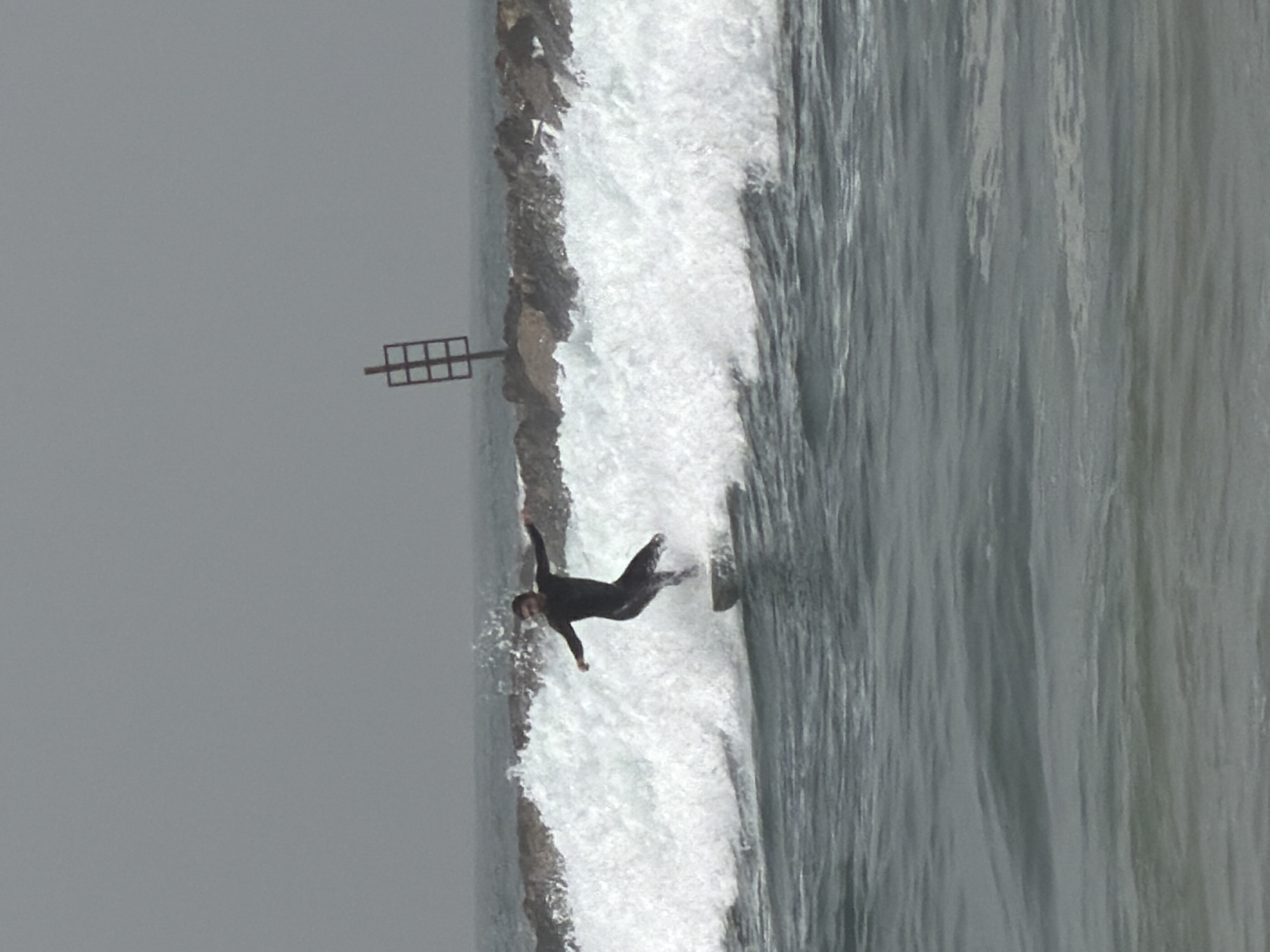 A sideways view shows a person in a black wetsuit, appearing to have fallen from the rocks, now partially submerged in the white water of the surf. The location is along the shoreline, presumably in the city of Tel Aviv, Israel. A metal frame is anchored along the shore, next to a rock outcrop, and the sea extends out into the distance under a gray sky.