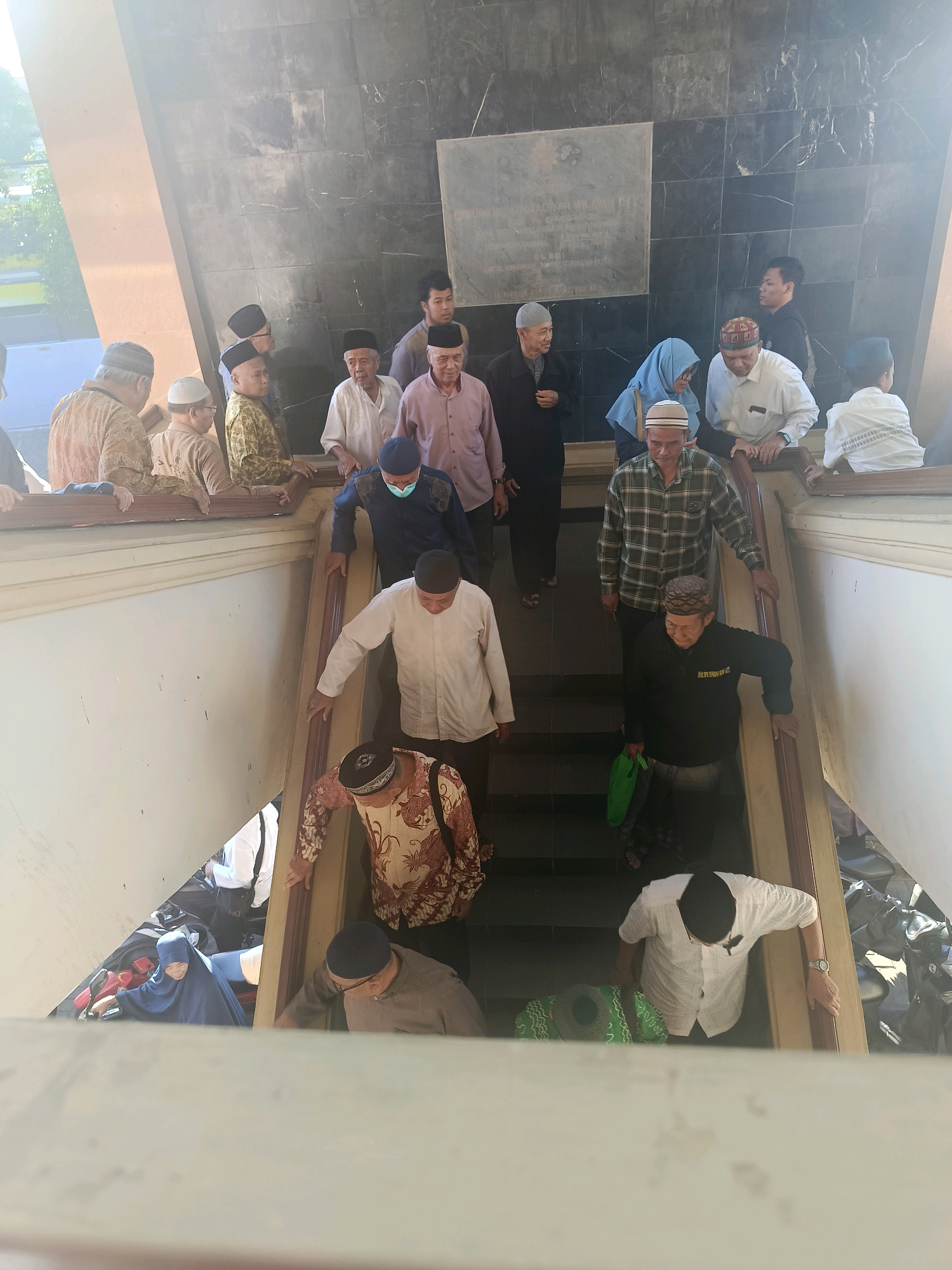 The image depicts a group of people, primarily men wearing traditional Islamic attire including peci hats and sarongs, descending a wide, light-colored staircase. Some individuals are dressed in white shirts, while others wear patterned or collared shirts. One man in the foreground wears a black long-sleeved shirt with text and a patterned sarong, carrying a green bag. Several people are looking down or engaged in conversation. The staircase is flanked by wooden railings, and the background features a dark, marbled wall with a plaque or inscription. The scene appears to be outdoors or in a well-lit, open-air structure, possibly a religious or historical site. The bright sunlight suggests a daytime setting. The presence of motorcycles parked in the background indicates a location within a city. Based on the style of dress, architecture, and the overall atmosphere, this scene could plausibly be located in Yogyakarta, Indonesia, a city known for its rich cultural heritage and Islamic traditions.