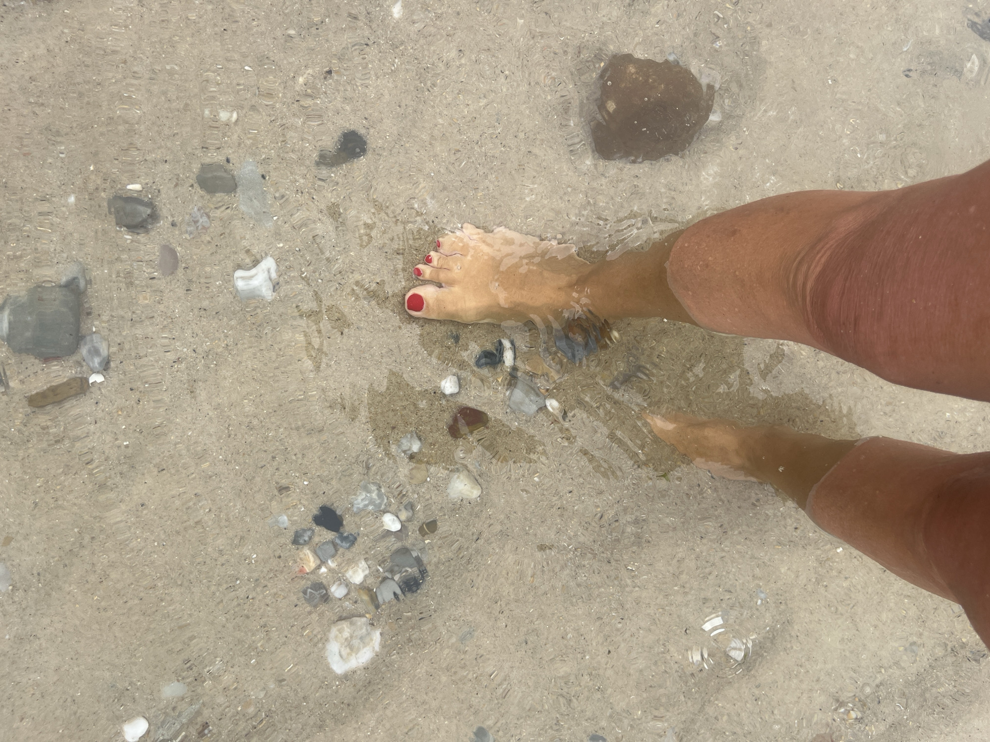 The image shows a person's legs and feet submerged in shallow water at a beach. The water is clear, revealing the sandy bottom and numerous small stones. The person's feet are bare, with toenails painted red. The water creates ripples and distortions on the surface. The scene depicts a person walking in the sea. This is likely the beach of Foz da Areia, located in the municipality of Caldas da Rainha, Portugal.