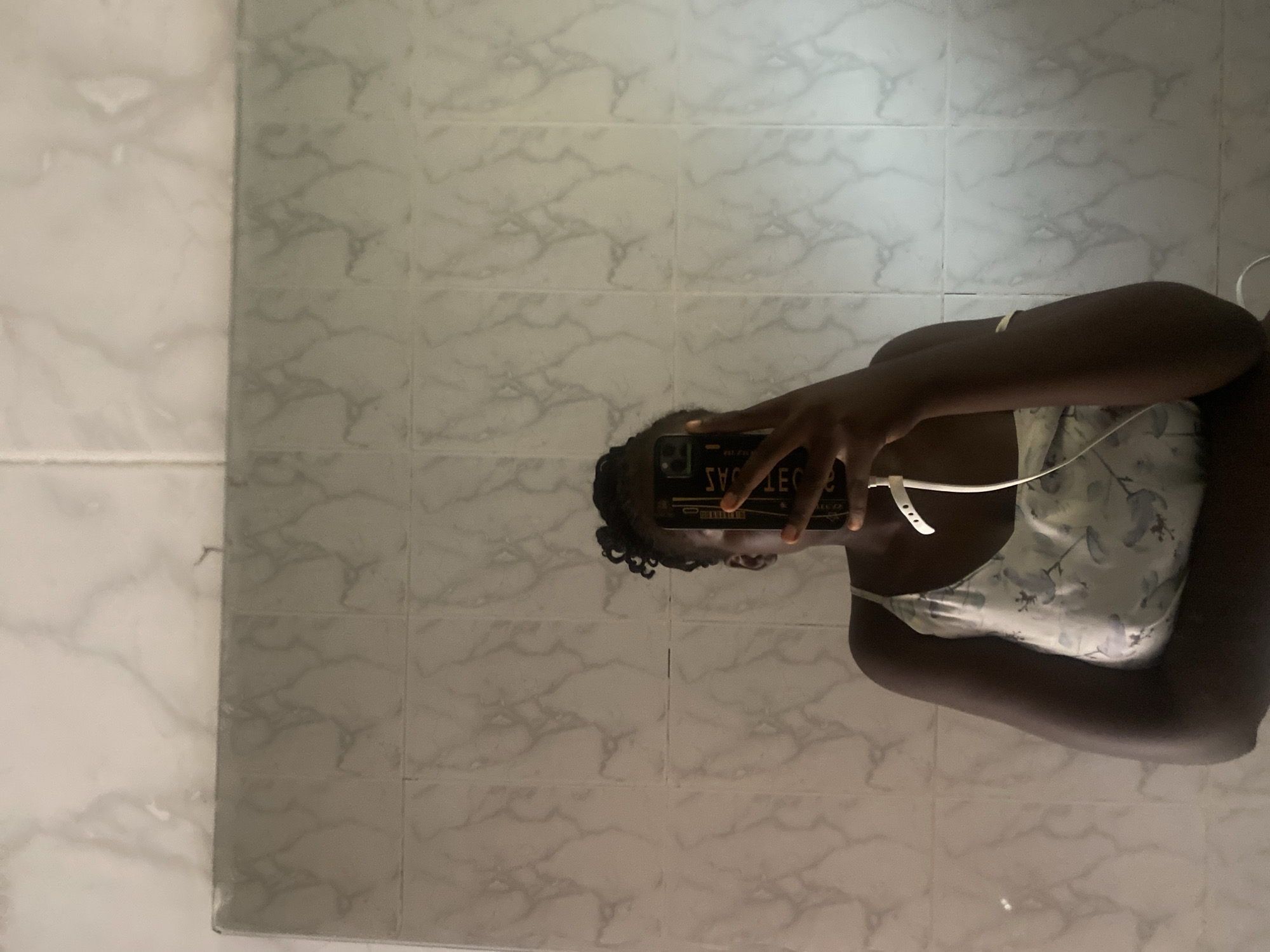 The image shows a young woman taking a mirror selfie in what appears to be a bathroom or tiled room. She is holding a smartphone to capture the reflection of herself. The background consists of tiled walls with a marble-like pattern. She is wearing a light-colored, floral-patterned camisole or tank top. Her hair is dark and styled in curls. The lighting suggests it could be daytime, possibly indoors with artificial light. The reflection of the phone shows text which is reversed due to the mirror, and it's difficult to discern clearly but appears to have some writing. The overall mood is casual and personal, as is typical of a selfie. There are no obvious indicators of weather or specific time of day beyond general indoor lighting. The location context provided is Wauro Leggal, Nigeria.