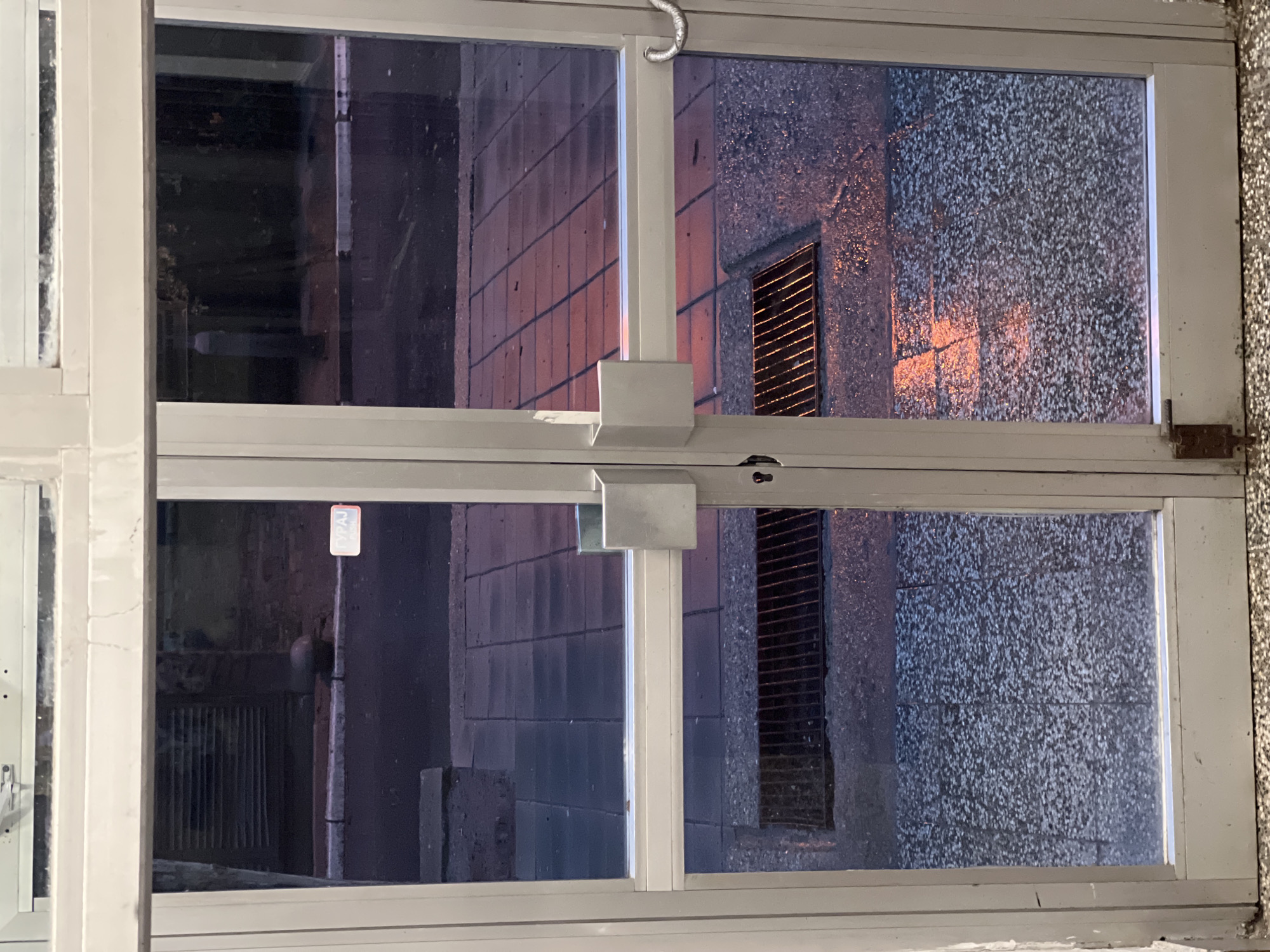 An interior view through a partially open, white-framed window reveals the exterior urban environment. The window is divided into four panes by a horizontal and vertical frame. The building exterior visible through the right panes is a textured wall with a dark, vertical grate-like structure. The left panes show a view down an alleyway. A street sign is visible in the distance. This is located in the city of Belgrade, Serbia.