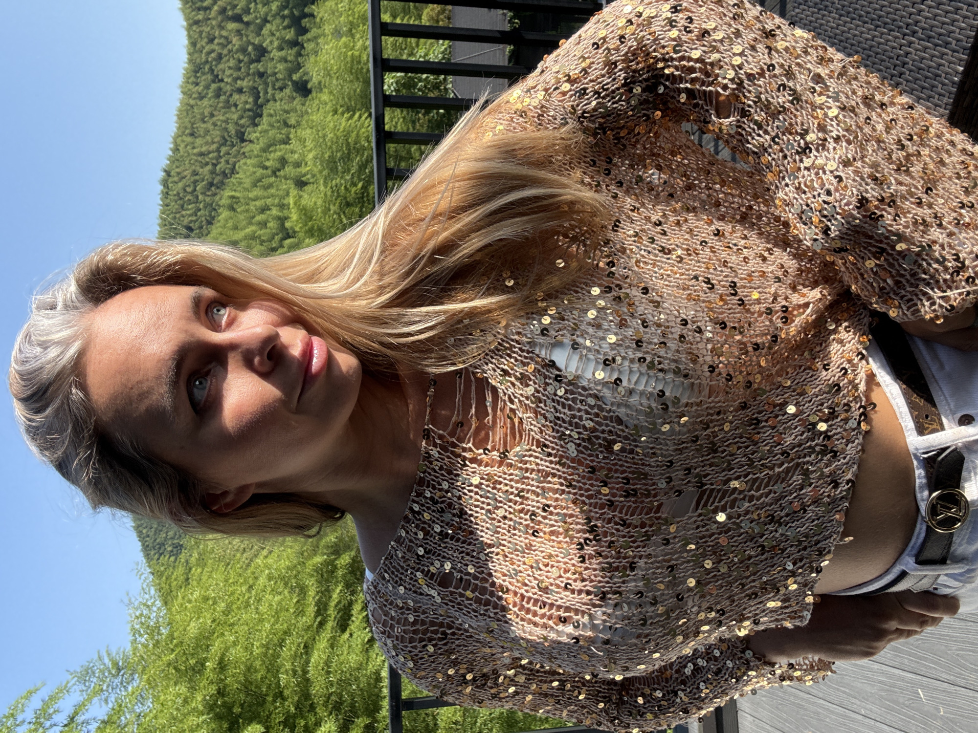 A blonde woman stands on a wooden deck, her body angled to the left, looking off-camera to the right. She wears a sheer, sequined, long-sleeved top over a white bra, and light-colored pants with a Louis Vuitton belt. Her hair is blonde, with some strands falling over her shoulder. Behind her, a black metal railing separates the deck from a background of lush green foliage and rolling hills, indicative of a natural, possibly mountainous, setting. The sky is clear blue. The lighting suggests it is daytime, possibly late afternoon, with sunlight casting shadows and highlighting the sequins on her top. This scene is not located in Wuxing District, China.