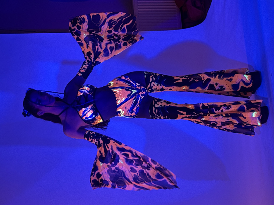 A full-body image captured indoors under blue monochromatic lighting reveals a person, likely female, standing centrally against a plain blue background. The individual is positioned with arms extended outwards and slightly downwards, head slightly tilted to the right. They are wearing a three-piece outfit with a black base and a prominent, glowing orange and yellow abstract pattern. This ensemble consists of a halter-neck crop top with a laced-up front, detached bell-sleeves that flare at the wrists, and high-waisted, flared pants with thigh cut-outs. Dark, possibly platform, footwear is visible beneath the pant hems. The subject's dark hair is styled with two small buns or knots on top, and subtle glowing elements are visible on the face. To the far right of the frame, a white vertical radiator and a partial red object are visible against the blue wall. The scene is located within a room in Sofia, Bulgaria.