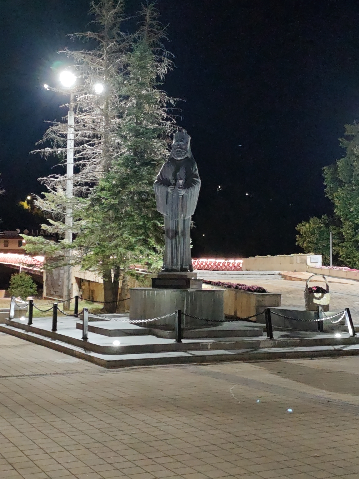 The image depicts a nighttime scene featuring a statue of a robed figure, likely religious in nature, standing on a tiered stone pedestal. The statue is illuminated by artificial lights. A lamppost with two bright lights is visible to the left of the statue, partially obscured by a tall evergreen tree. The area around the pedestal is cordoned off by metal chains and posts. In the foreground, a paved area with a diamond pattern is visible. In the background, there are buildings with illuminated windows, a wall with decorative red and white elements, and trees. A basket filled with flowers is placed to the right of the pedestal. The overall setting appears to be a public space, possibly a park or a square in the city of Lovech, Bulgaria, given the context.