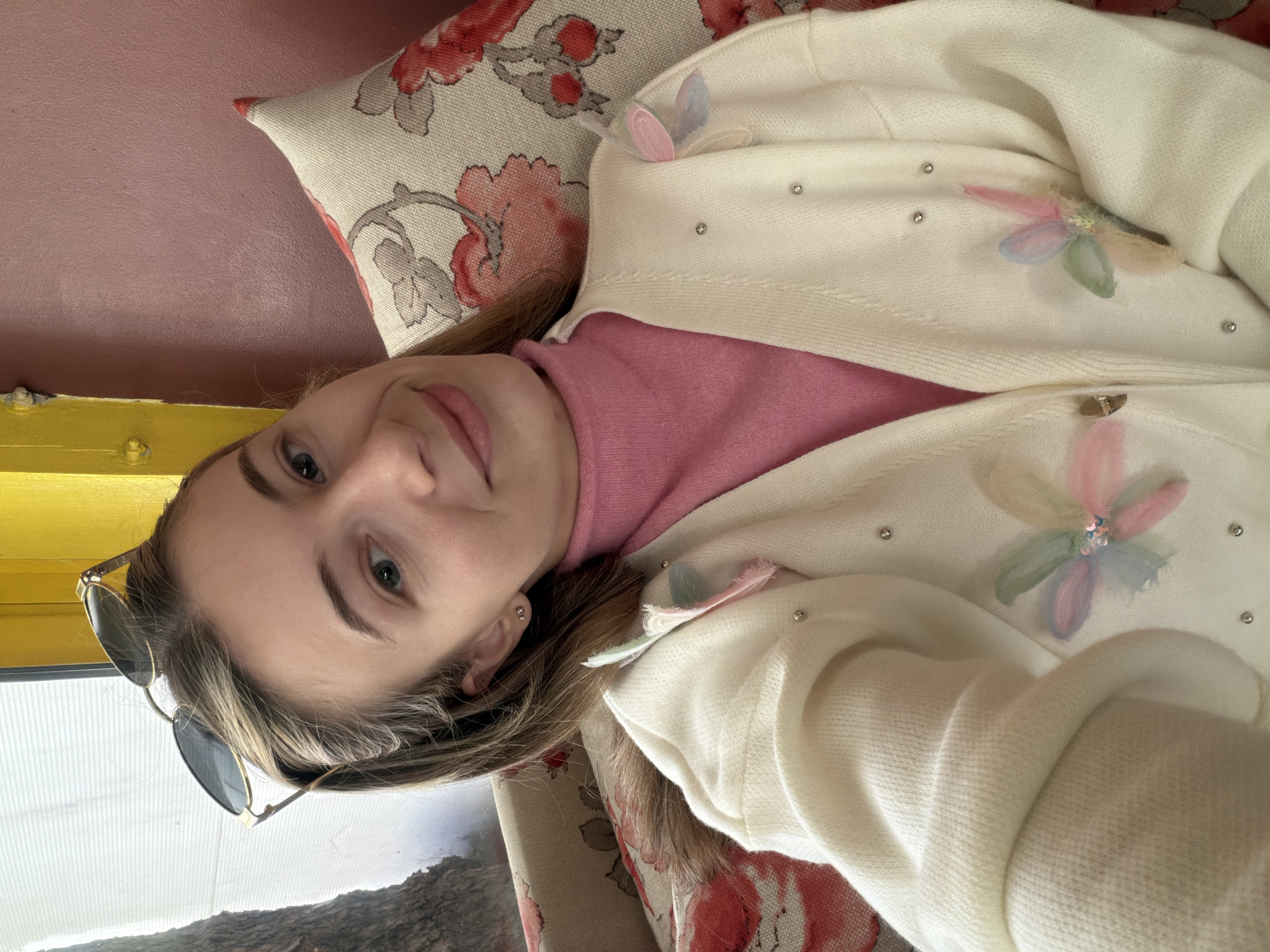 A woman with light brown hair and blue eyes is lying down, angled towards the camera. She wears a pink turtleneck sweater and a white cardigan decorated with fabric flowers and small studs. Sunglasses are perched on her head. She is resting on a patterned cushion with a floral design in shades of red and gray. The background shows a glimpse of a bright yellow wall. Through a window, there is a view of a mountainous landscape. The location is Plovdiv, Bulgaria, based on the geographical context.