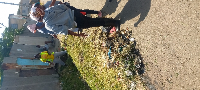 The image portrays an outdoor scene in an urban or peri-urban area of Nairobi, Kenya, under bright daylight. Five individuals are visible. In the mid-ground left, a person wearing a yellow t-shirt with a circular logo, a yellow safety vest, light grey pants, and dark footwear is actively using a stick to clear debris and vegetation from a grassy verge. To their right, three individuals are partially visible. One individual, facing away from the camera, wears a grey hooded sweatshirt, dark pants, and black rubber boots, holding a stick pointing towards the ground. Two additional individuals, wearing light blue tops and patterned head coverings, stand nearby, with one also holding a stick. In the background, a male individual dressed in a light-colored t-shirt and shorts is seated against a building structure, observing the activity. A significant accumulation of assorted refuse, including plastics, fabric, and organic material, is present on the grassy verge separating a dirt road from a tall, makeshift wall. This wall is constructed from corrugated metal sheets, some exhibiting rust, and features a light blue wooden door along with another darker door. Beyond the wall, residential-style buildings with light-colored walls and a partial blue roof are visible. The foreground consists of a dirt and gravel road, with clear shadows indicating strong sunlight.