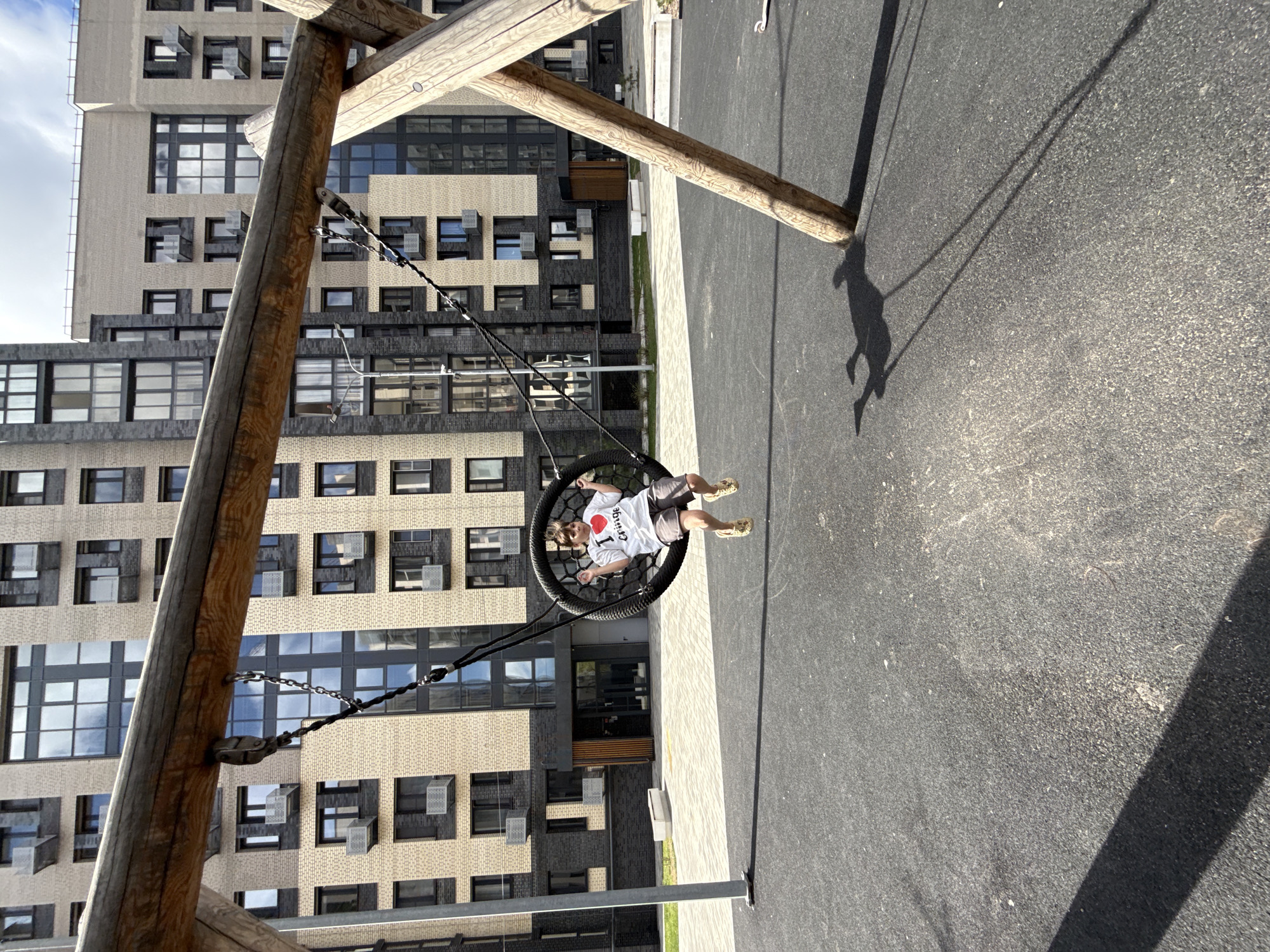 The image shows a child swinging in a basket swing in a playground. The child is wearing a white t-shirt with a red heart and the word "cringe" on it, shorts, and yellow sandals. The swing is attached to a wooden structure. Behind the swing is a modern apartment building with many windows. The playground surface is dark gray. The location appears to be an urban setting, specifically at Unknown, Unknown. The sun is casting a shadow of the swing and the child on the ground.