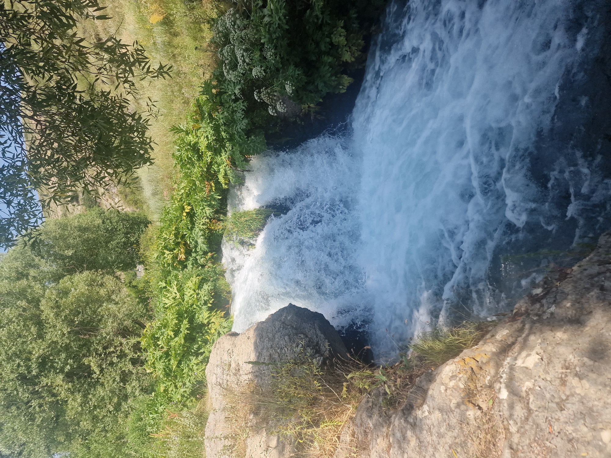 This media file depicts a powerful waterfall cascading down a rocky incline amidst lush green vegetation. The water is a vibrant, frothy white, indicative of its turbulent descent. Surrounding the waterfall are dense clusters of broad-leafed plants and trees, their leaves a rich green, suggesting a warm and sunny day. Sunlight dapples the scene, highlighting the textures of the rocks and the movement of the water. The overall impression is one of natural beauty and untamed energy. The time of day appears to be daytime, with ample sunlight illuminating the scene. The location is identified as Sisian, Armenia, which is known for its picturesque natural landscapes. No people, objects, or text are visible in the frame, focusing solely on the raw elements of nature.