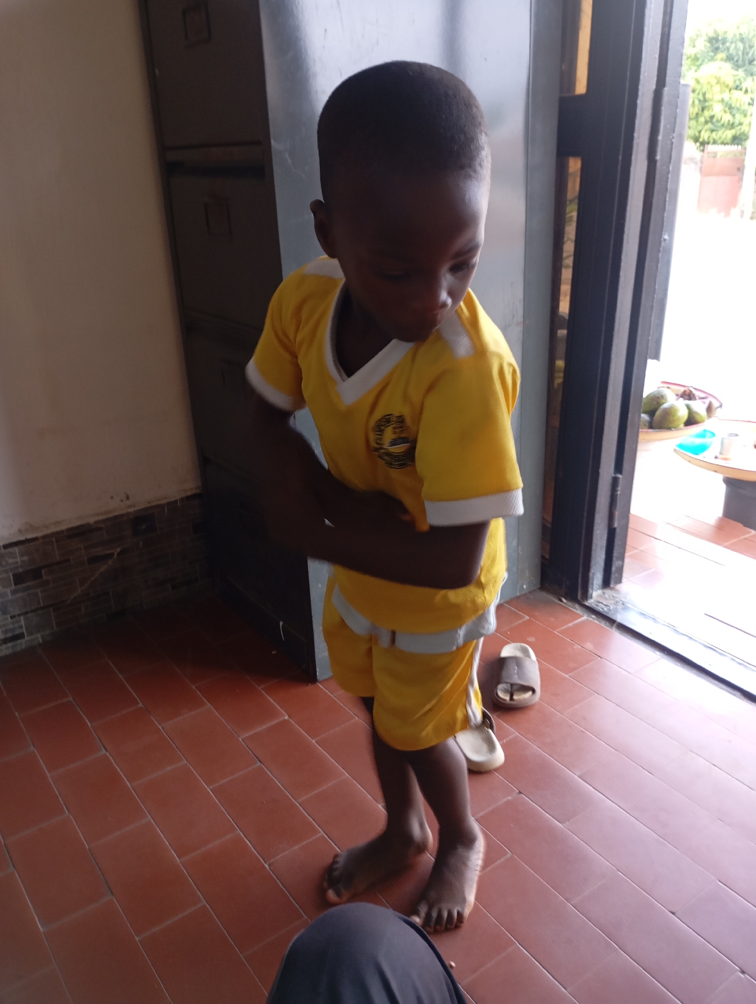 A young child, appearing to be between 3 and 5 years old, stands barefoot on a reddish-brown tiled floor. The child is wearing a yellow t-shirt with white trim around the collar and sleeves, and matching yellow shorts with white trim. The child's arms are crossed in front of their body, and they are looking down. To the right of the child, on the floor, are two open-toed sandals, one light brown and one beige. In the background, to the left, is a tall, grey metal cabinet with visible handles and indentations. To the right, a doorway or opening reveals an outdoor scene with greenery and what appears to be a table with fruit. The lighting suggests an indoor setting near an entrance. This scene is located in Jos, Nigeria.