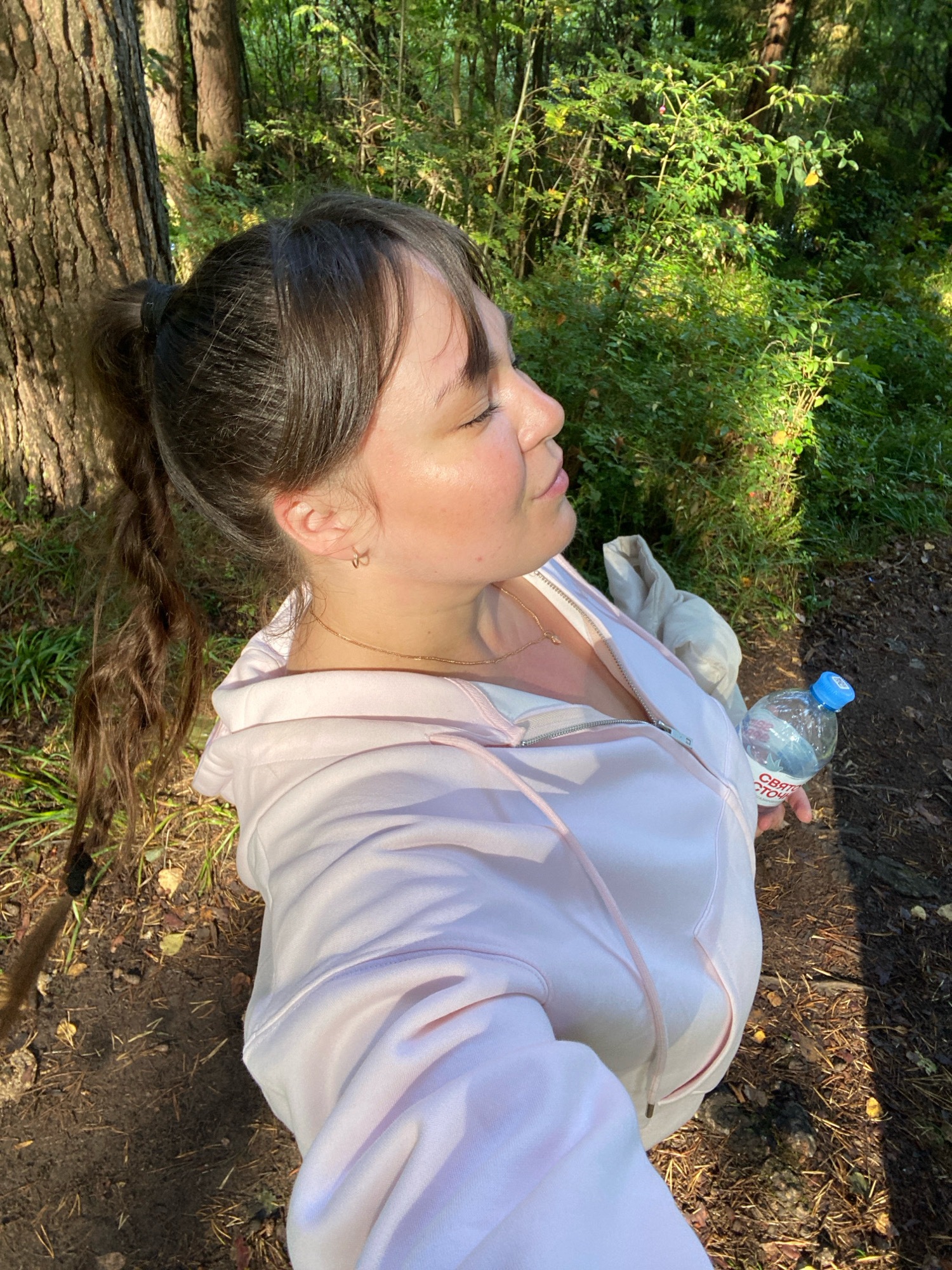 The image shows a young woman in a light pink hooded jacket standing in a forest. The sun is shining, casting dappled shadows on the ground. The woman has her eyes closed, and her face is tilted upwards, as if enjoying the sunlight. Her dark brown hair is pulled back into a ponytail. She is holding a plastic bottle of water in her right hand. The label on the bottle has Russian text, which includes the words "Свято" and "СТО". The background is filled with lush green foliage and trees, typical of a forest setting. The ground is covered with dirt, fallen leaves, and pine needles. The overall impression is one of peace and relaxation in nature.