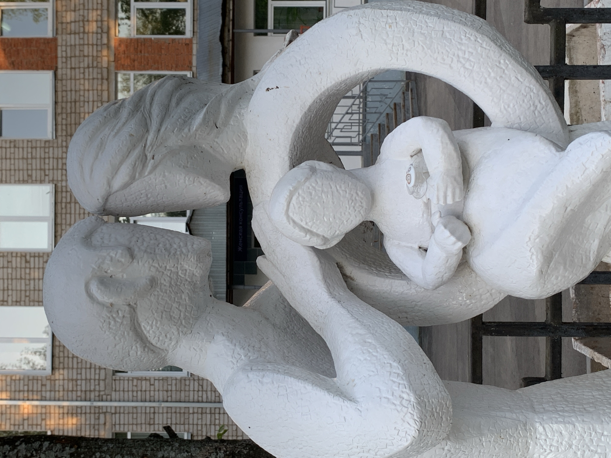 A white, abstract sculpture of a man, woman, and baby is outdoors in front of a building. The man, on the left, looks at the woman in the center, who holds a baby above her. The woman and baby are encircled by a large, textured ring. The building behind the sculpture is brick on one side with white windows, and the other side is white with a gray staircase visible through a doorway. The sculpture is located in the city of Chaikovsky, Russia.