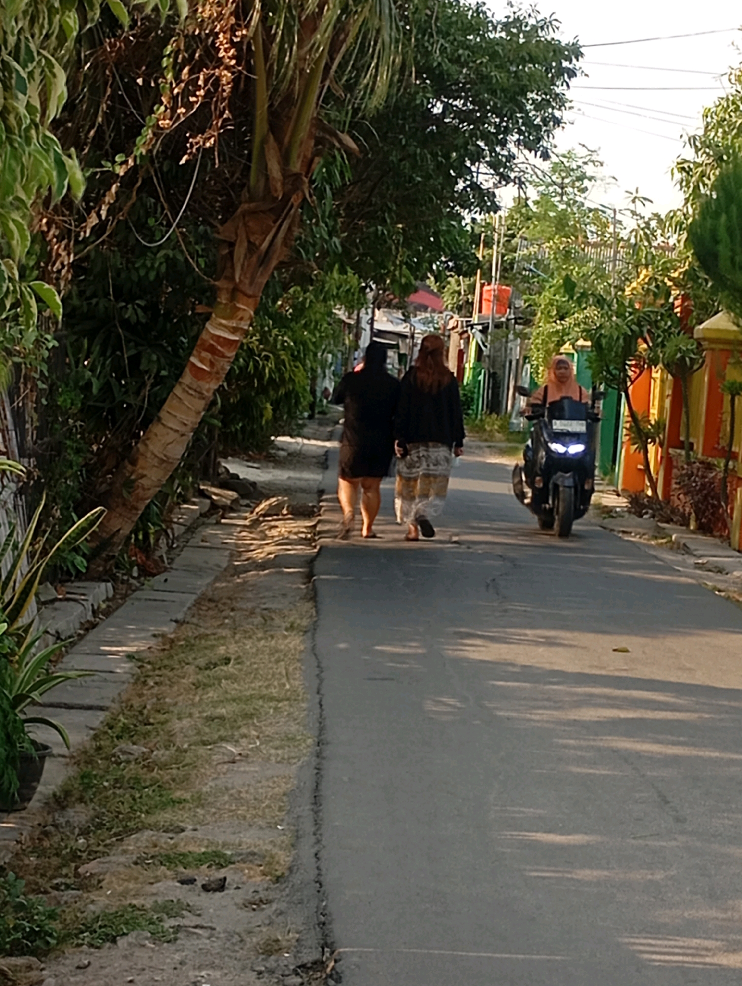 The image depicts a street scene in Babelan, Indonesia, bathed in the warm light of late afternoon or early morning. Two individuals are walking away from the camera on the right side of the asphalt road, their backs to the viewer. The person on the left wears a dark top and a patterned skirt, while the person on the right wears a black top and a patterned skirt. Further down the road, a person wearing a headscarf rides a dark-colored motorcycle, its headlights illuminated. The road is bordered by a sidewalk on the left, with vegetation, including a palm tree, growing along its edge. Behind the sidewalk, a low wall and houses with brightly colored walls are visible. The right side of the road features more houses with colorful facades and some trees. Sunlight casts long shadows across the road.