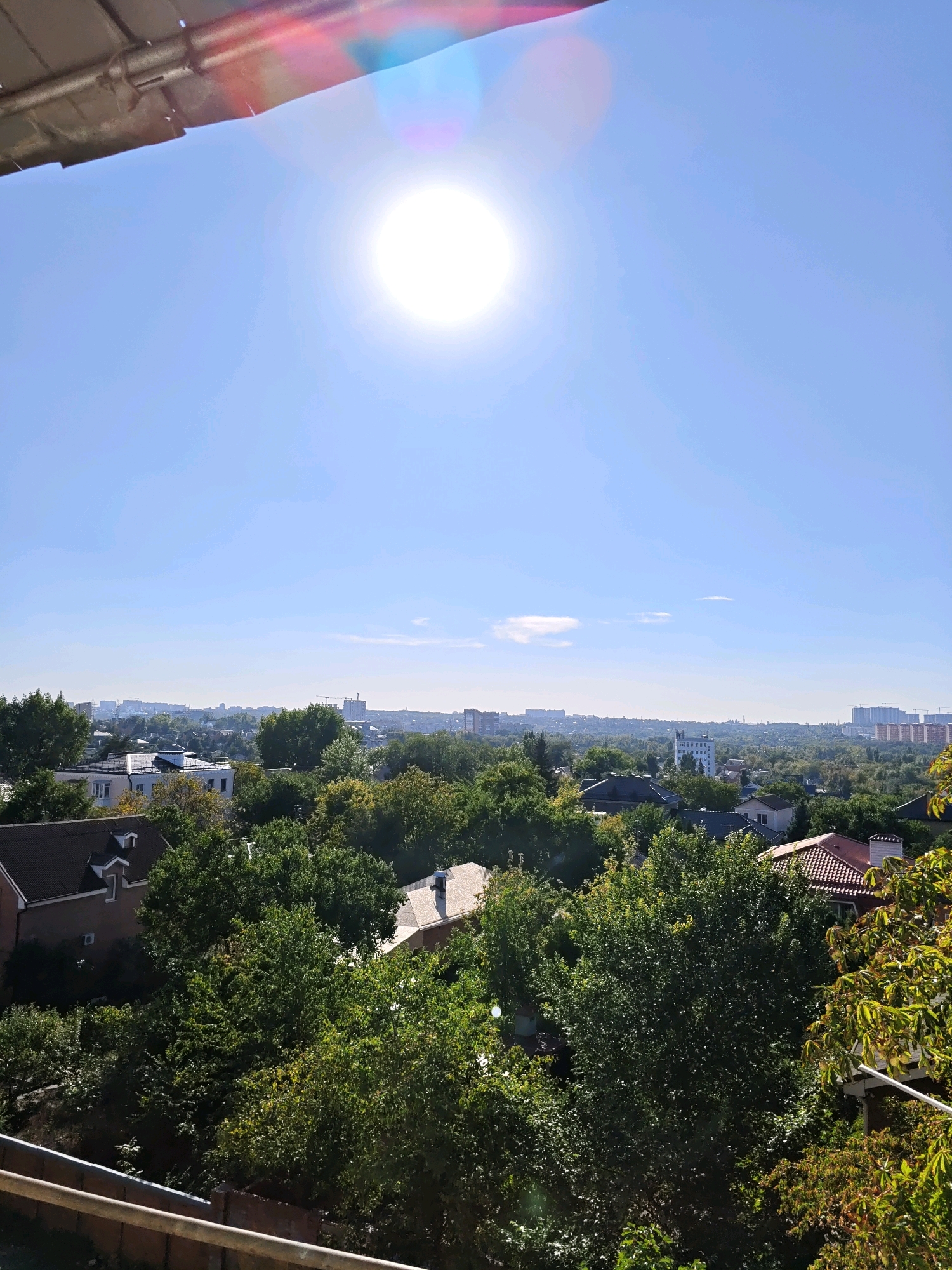 The image depicts a bright, sunny day in Rostov-on-Don, Russia. The sun is high in the sky, casting a strong glare with lens flare effects. Below the sky, a dense canopy of green trees fills the midground, interspersed with the rooftops of residential buildings. The trees vary in shades of green, suggesting a mix of deciduous and possibly evergreen species. Several houses with pitched roofs are visible, some with brick exteriors and others with lighter colored siding. In the background, a distant cityscape with various buildings, including some taller structures and cranes, stretches across the horizon. The foreground features a portion of a metallic railing and what appears to be a wooden fence, suggesting the viewpoint is from a balcony or elevated outdoor space. The overall scene conveys a residential urban environment on a clear day.
