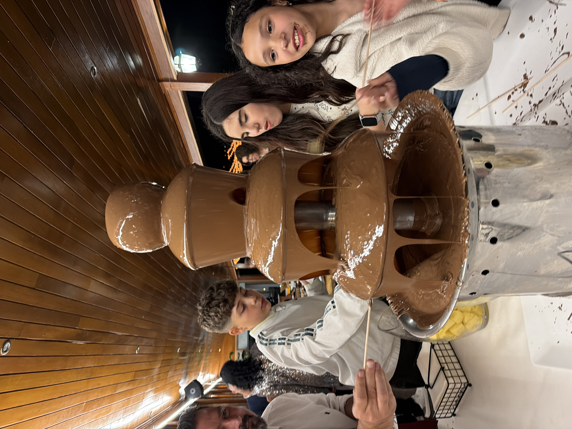 A group of people, including two children, are standing around a chocolate fountain in Vila Real, Portugal. The fountain is made of stainless steel and has four tiers. The chocolate is flowing smoothly. The children appear to be enjoying the fountain. The background shows what looks to be the inside of a wooden building.