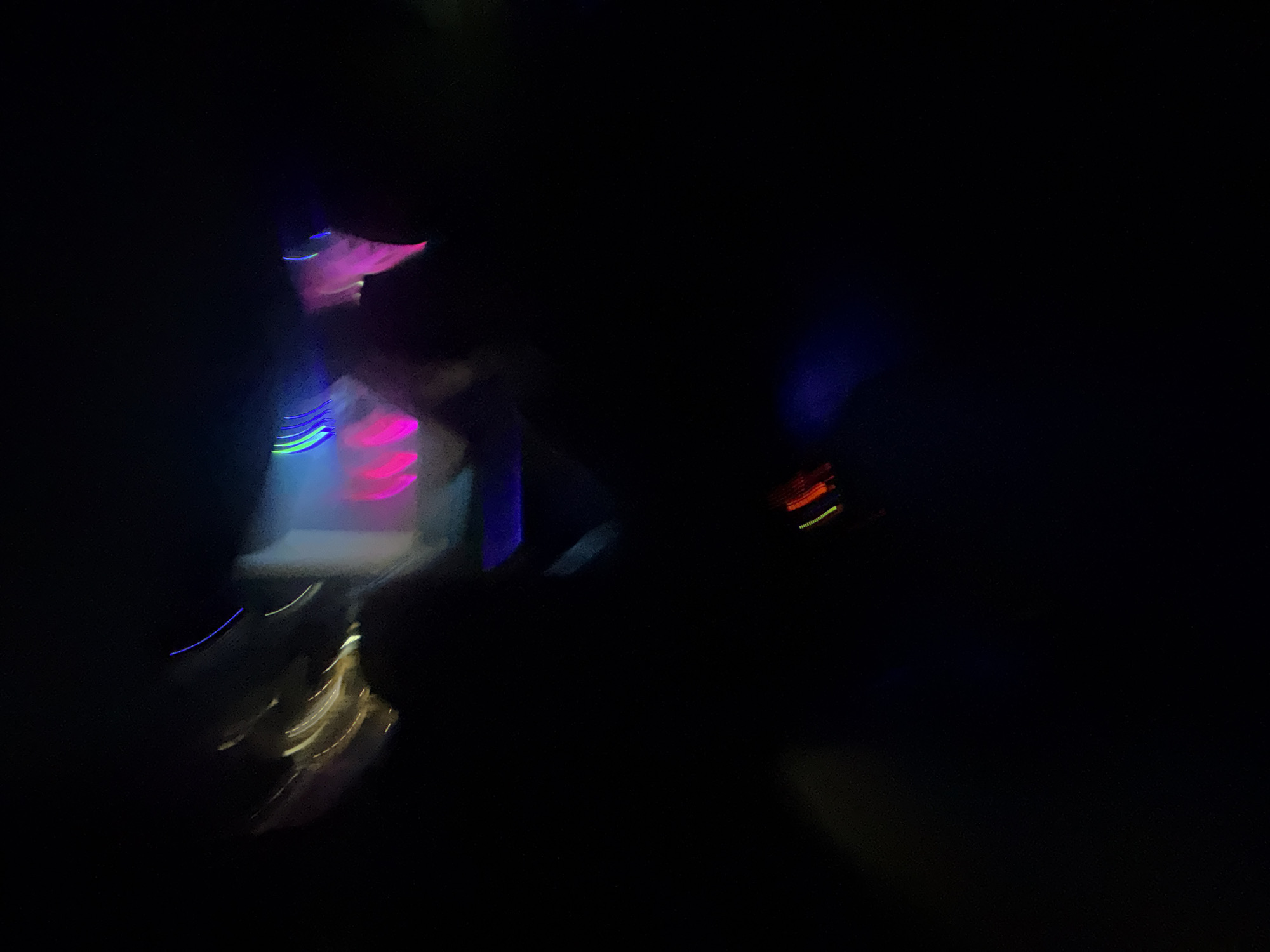 The image, taken in San Bernardino Tlaxcalancingo, Mexico, is blurry and dark. There are streaks of light in pink, green, blue, orange, and yellow. There is no identifiable person, subject, or object. Interactions are indeterminable due to the blur. The environment is dark.