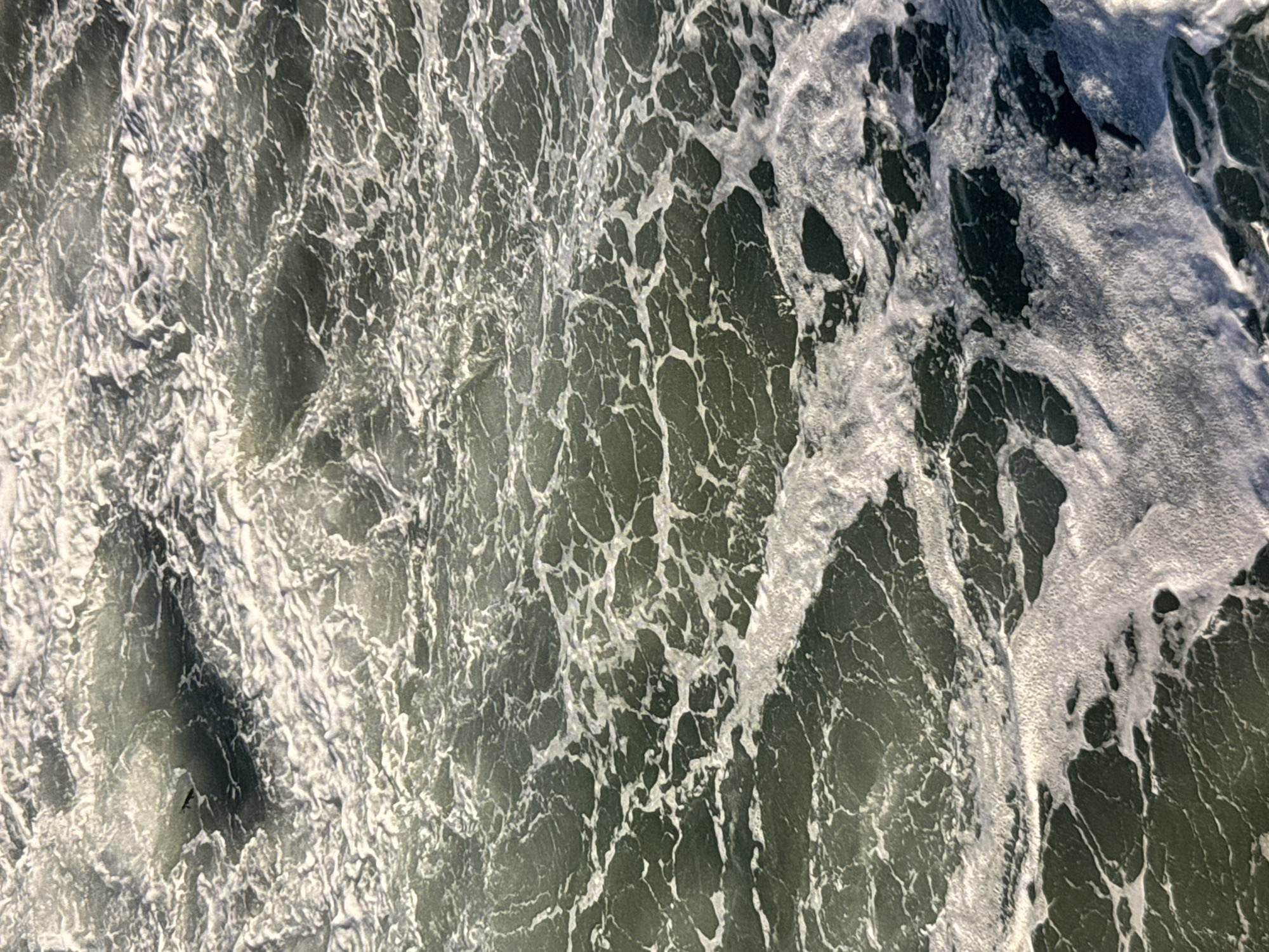 This is a close-up, high-angle shot of choppy, dark green ocean water with white foam and waves. The water is textured with swirling patterns of foam, resembling a web or a cracked surface. Some darker patches of water suggest deeper areas or shadows. The overall impression is of a dynamic and turbulent sea. The image is an abstract representation of the ocean, likely taken at a beach in Los Angeles, United States of America. There are no people, specific landmarks, or identifiable objects visible in the frame, focusing solely on the natural patterns of the water.
