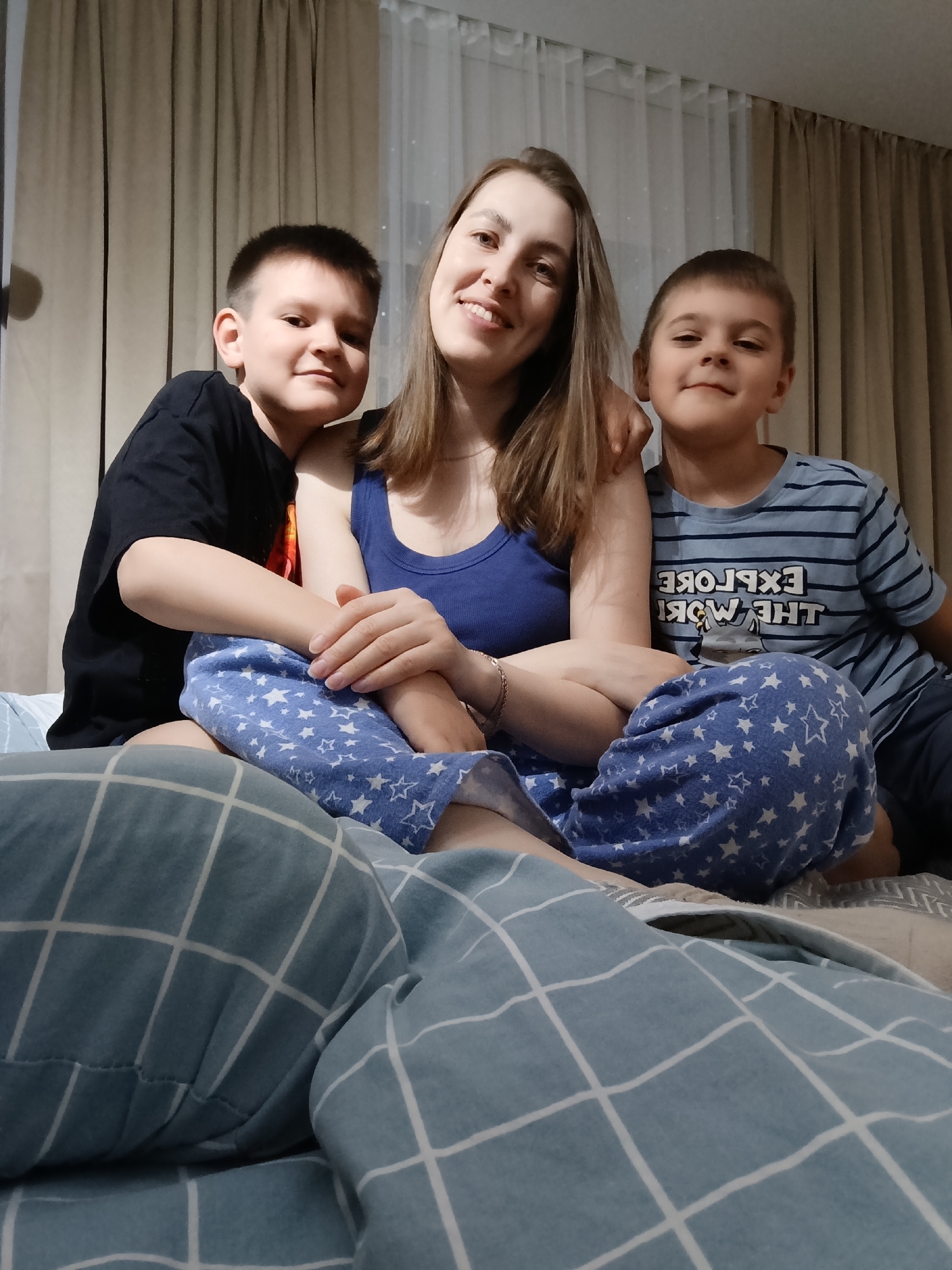 This image depicts three individuals, one adult female and two male children, seated in an indoor residential setting in Chaikovsky, Russia. The adult female is positioned centrally, wearing a blue tank top and blue pajama pants adorned with white star patterns. Her hands are clasped in her lap. To her left, a male child, estimated to be between 8 and 10 years old, wears a black t-shirt and has his left arm draped over the female's shoulder. To her right, a second male child, also estimated between 8 and 10 years old, wears a light blue striped t-shirt featuring the inverted text "EXPLORE THE WORLD" and has his right arm over the female's shoulder. All three subjects are smiling and looking directly at the camera. They are seated on what appears to be a bed, with a light blue and white checkered blanket or duvet partially visible in the foreground. The background reveals light brown curtains covering a window, with sheer white curtains behind them. The scene is illuminated by ambient indoor light.