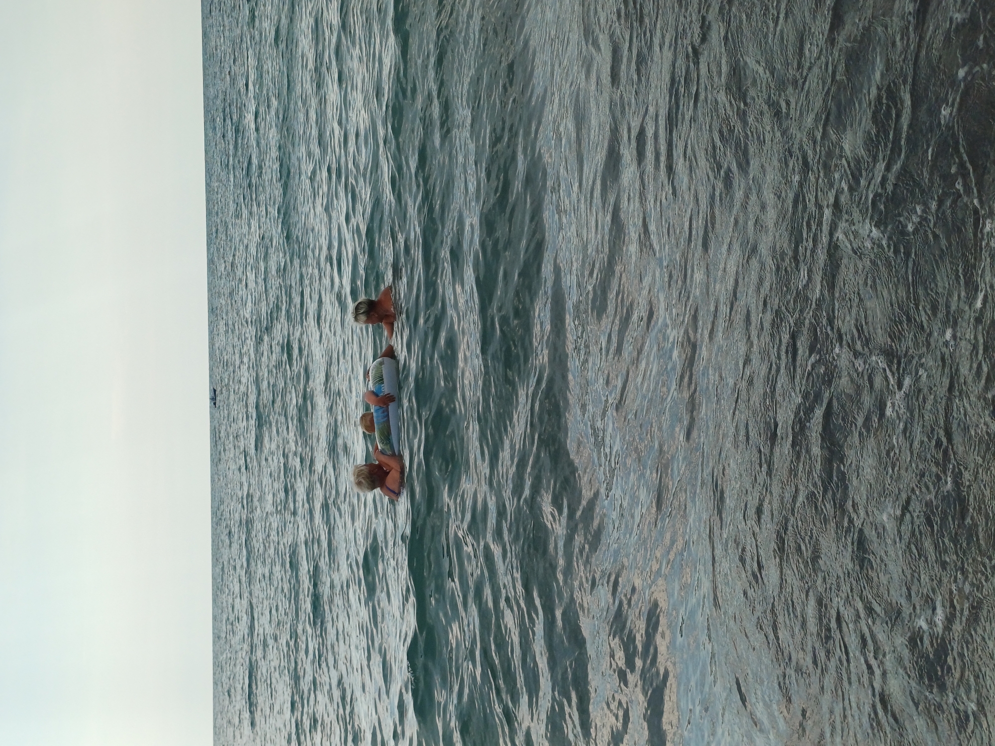 The image shows three people in the ocean. The person in the foreground is a child with blond hair, wearing a blue swimsuit. They are lying on their back on a colorful inflatable raft. Behind them, another child with blond hair is partially visible, also on the raft. Further back, another person, possibly an adult, is in the water, with their head and shoulders visible. The water is a deep blue-green color, with ripples and small waves. The sky is a pale, overcast gray. The scene appears to be a calm day at the beach. The sun is not strongly visible, suggesting it might be early morning or late afternoon. There is no visible text in the image.