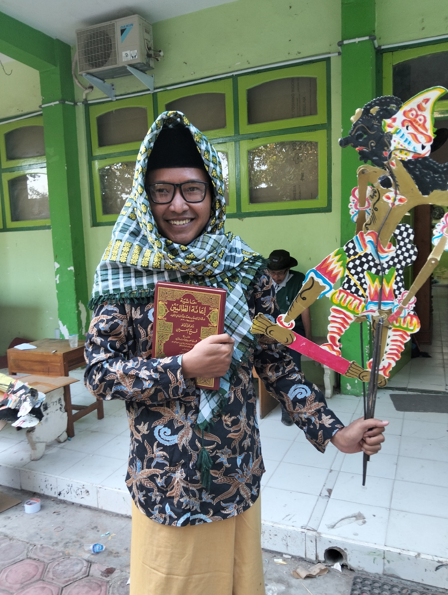 In a courtyard, a man wearing a black peci hat, glasses, a patterned batik shirt, and a plaid sarong, stands smiling. He holds a red book with Arabic script in his left hand and a wayang golek puppet in his right. The background shows a green building with windows, an air conditioning unit, and a wooden table. The ground is tiled, with some debris. The image appears to be taken in Kranji, Indonesia.