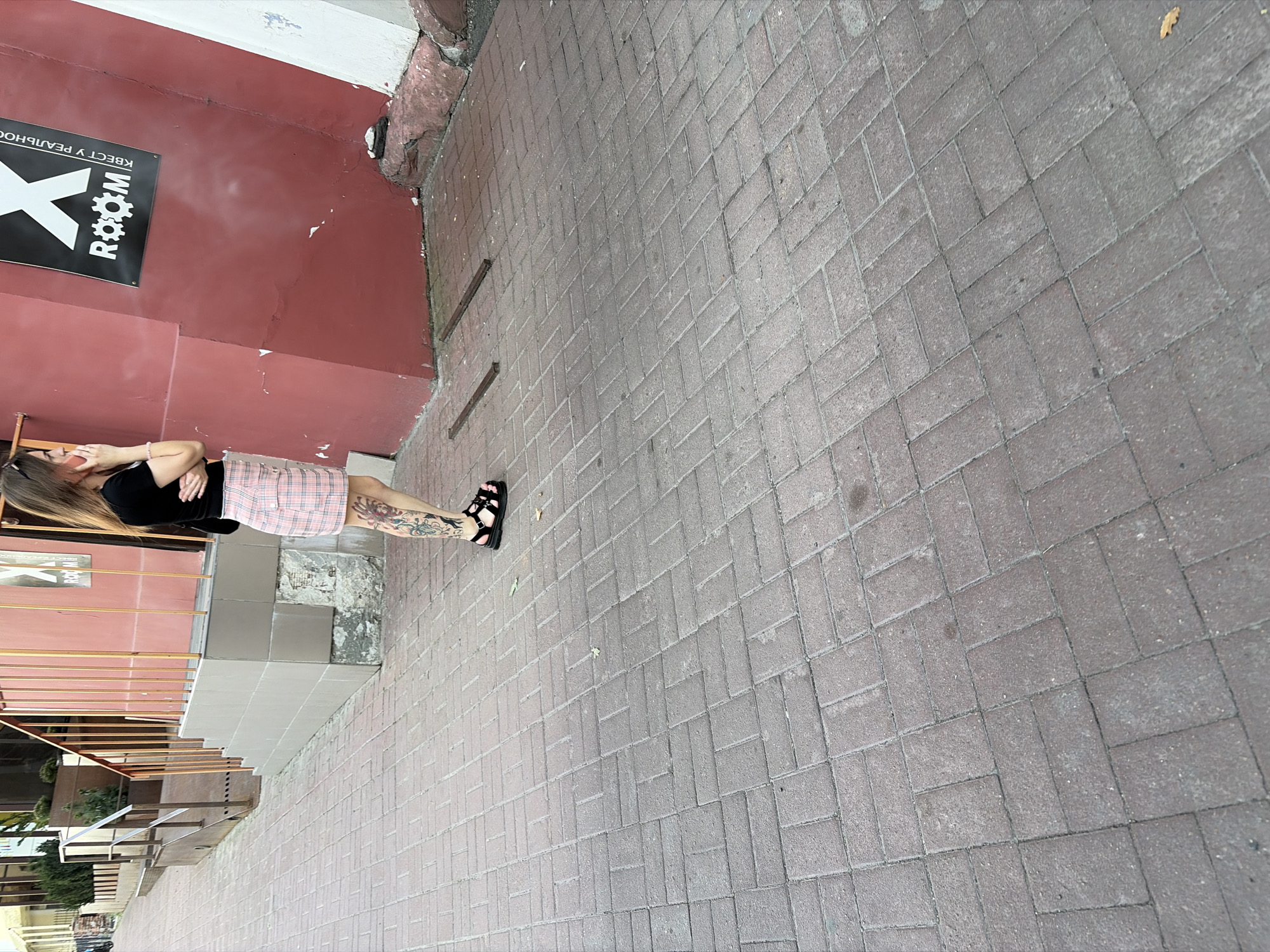 A woman with long blonde hair, wearing a black t-shirt and a pink plaid skirt, stands on a brick-paved sidewalk in Chernihiv, Ukraine. She is holding a pink mobile phone to her right ear with her right hand, and her left hand is crossed over her torso. The woman has tattoos on her left leg, which is visible below her skirt. She is wearing black sandals with buckles. Behind her, to her right, is a red wall with a black sign that reads "X ROOM" in white letters. To her left, there is a metal railing and a building facade with a set of stairs. The sidewalk is made of reddish-brown bricks laid in a herringbone pattern.