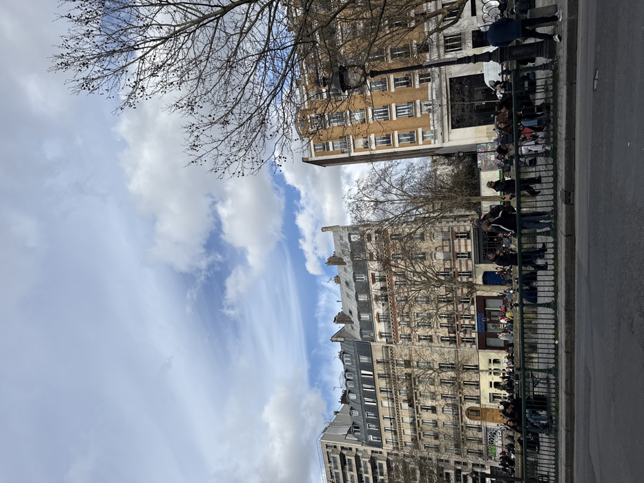 This image depicts an urban scene along the Canal Saint-Martin in Paris, France, likely captured during late winter or early spring based on the bare trees.
In the upper portion of the frame, a partly cloudy sky shows patches of blue interspersed with white, wispy clouds. The background features a row of multi-story Parisian buildings. On the far left, a modern, light-colored apartment building with numerous balconies is visible. Adjacent to it are two classic Haussmannian-style buildings, characterized by cream-colored facades, multiple windows with small iron balconies, and grey mansard roofs with dormer windows. The ground floors of these buildings appear to house commercial establishments. To the right of these, a dense cluster of bare deciduous trees stands, and further right, a taller, distinct building with a light brown facade and multiple small balconies is prominent. An ornate black lamppost stands in front of this building.
The midground is dominated by a public space alongside a waterway, separated from the viewer's immediate foreground by a dark green, ornate metal railing. Beyond this railing, a significant number of people, approximately fifty to seventy individuals, are gathered. Many are seated directly on the ground or low ledges along the canal bank, while others stand or walk. They are dressed in casual outerwear, including jackets and coats, consistent with cool weather. Their interactions appear social, with groups conversing and relaxing. A white van is partially visible behind the group on the right.
The immediate foreground shows a dark grey asphalt road surface.