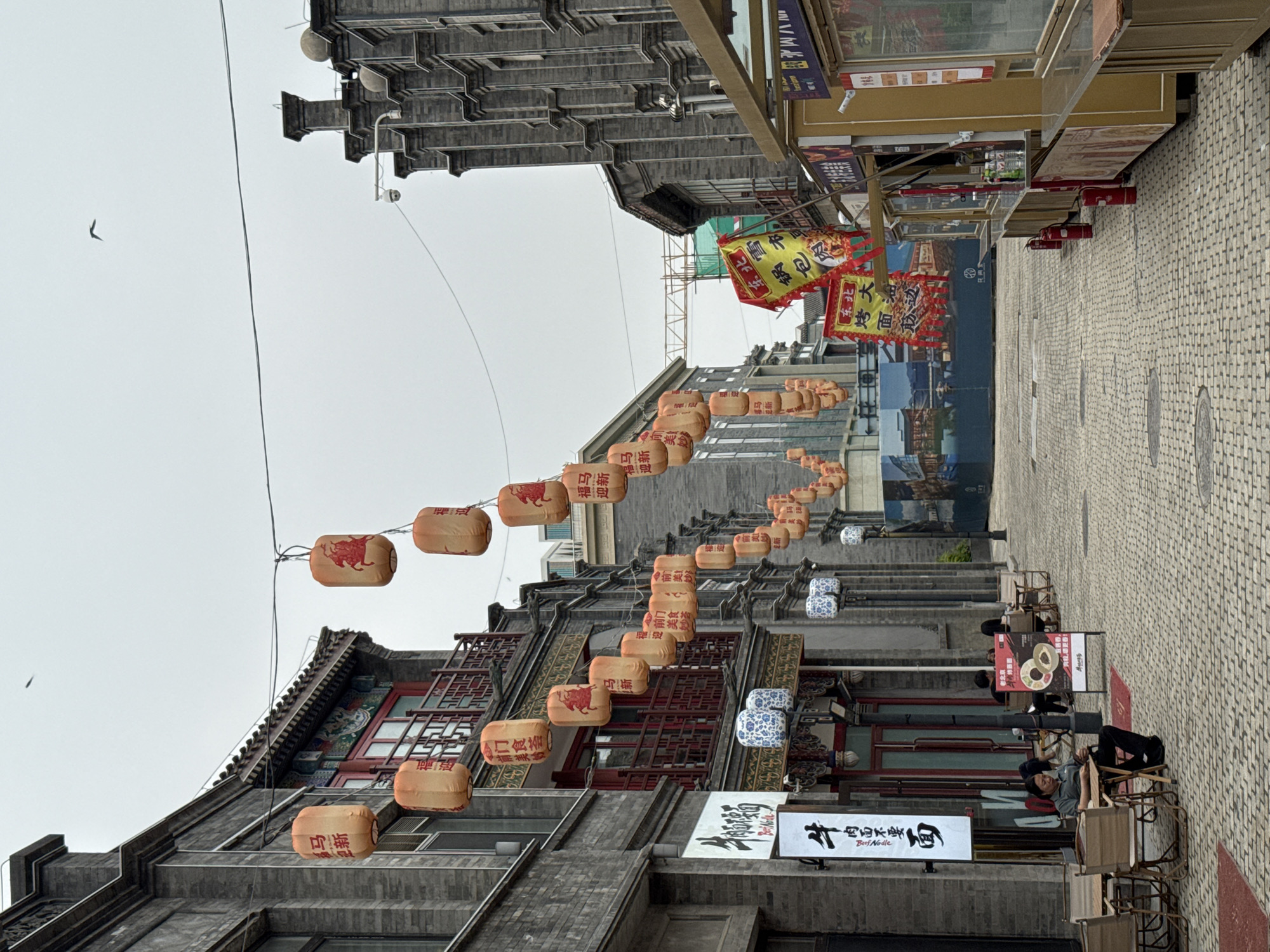 The image depicts a street scene in the Dongcheng District of Beijing, China. Traditional grey brick buildings line the street, adorned with strings of orange lanterns featuring Chinese characters and depictions of bulls, likely for a Lunar New Year celebration. Banners advertising local cuisine, such as "Guobaorou" and "Beef Noodles," are visible. People are seated in chairs along the sidewalk. The street is paved with cobblestones. The sky is overcast. A bird is flying in the upper left of the image.