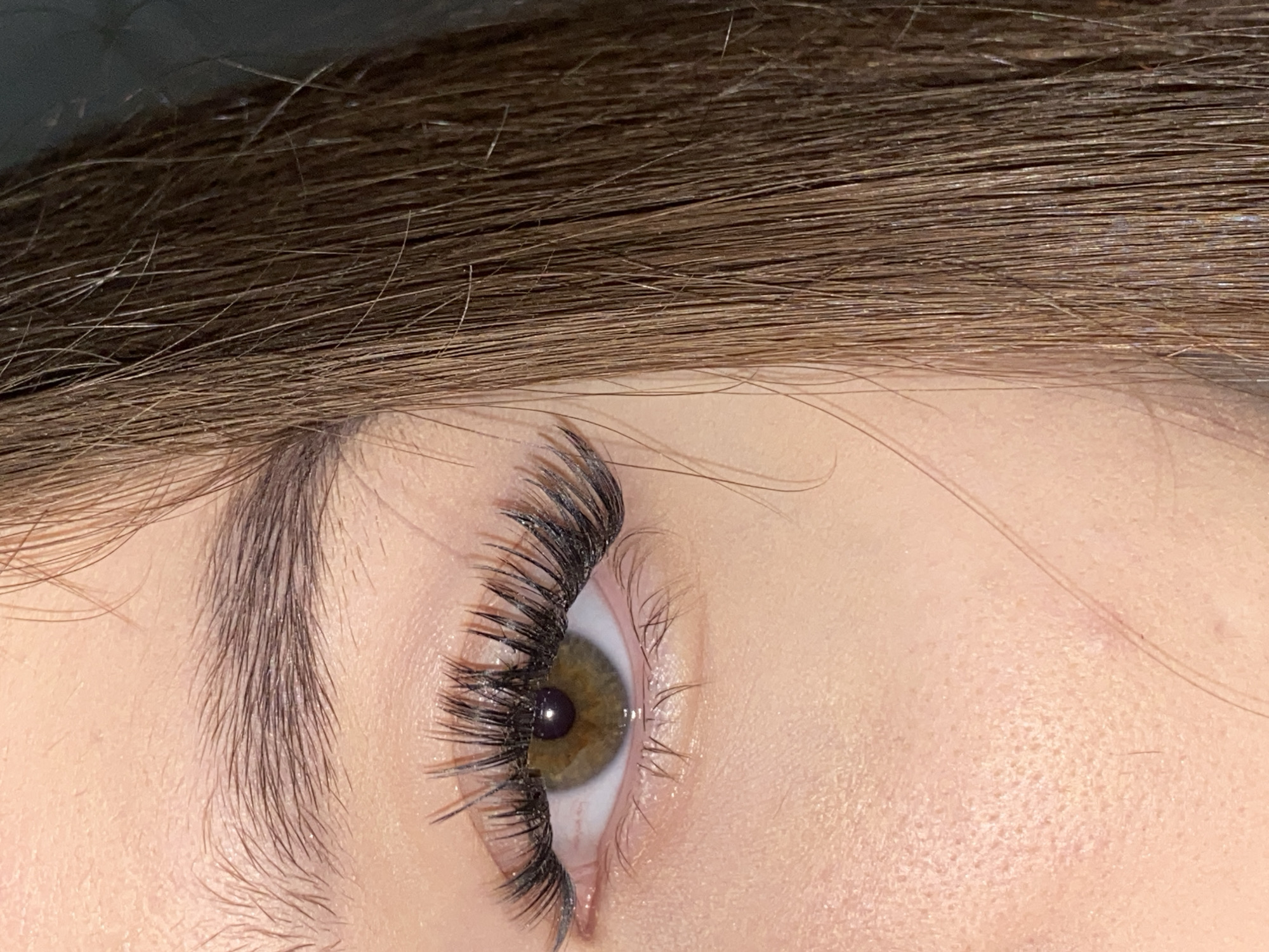 The image displays a close-up, vertical perspective of a human right eye and the surrounding facial area. The eye features a hazel iris, a dark pupil, and a prominent set of dark, volumized upper eyelashes, appearing to be lash extensions or heavily mascaraed natural lashes. The lower lashes are present but less pronounced. Above the eye, a dark brown, well-groomed eyebrow is visible. The skin visible on the cheek and forehead areas is light-toned with visible pores and minor texture. To the right side of the frame, strands of long, straight brown hair partially obscure the face, extending downwards. The background behind the hair is dark and indistinct. No specific actions, interactions, or environmental details are discernible. The location in the city of Chaikovsky, Russia, cannot be determined from the visual information provided.