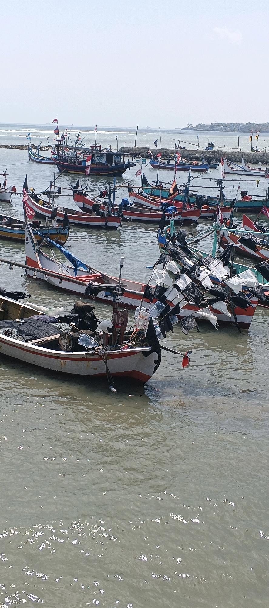 In Kandangsemangkon, Indonesia, a fleet of small fishing boats is moored in a harbor under a hazy sky. The boats, primarily made of wood, are painted in various colors, with many featuring red and white stripes along their hulls. Several boats are adorned with Indonesian flags and other colorful pennants. Fishing nets, equipment, and other supplies are visible on deck in some of the boats. In the background, a breakwater extends into the sea, with more boats and structures visible further out. The water is calm and appears somewhat murky, reflecting the overcast conditions. The scene depicts a typical working harbor.