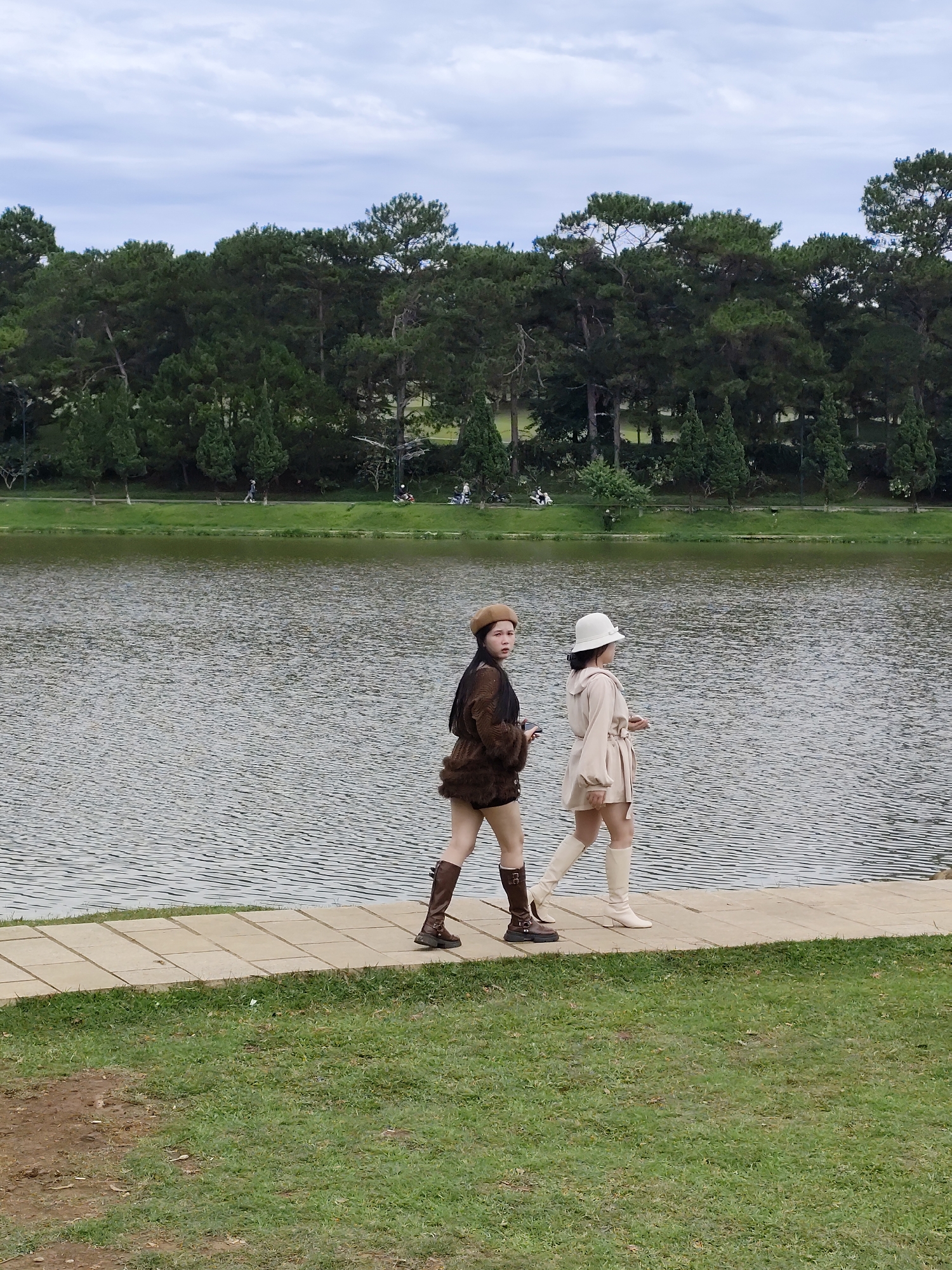 Two women are walking along a paved path next to a lake. The woman on the left has long black hair and wears a brown beret, a brown fuzzy jacket, shorts, and brown boots. The woman on the right wears a white hat, a light tan belted jacket, shorts, and cream-colored boots. Behind the path, there is a lake with trees along the opposite shore. In the distance, small figures of people can be seen along the shore. The path is bordered by green grass. The sky is cloudy. This is most likely Ho Xuan Huong Lake in Da Lat, Vietnam.