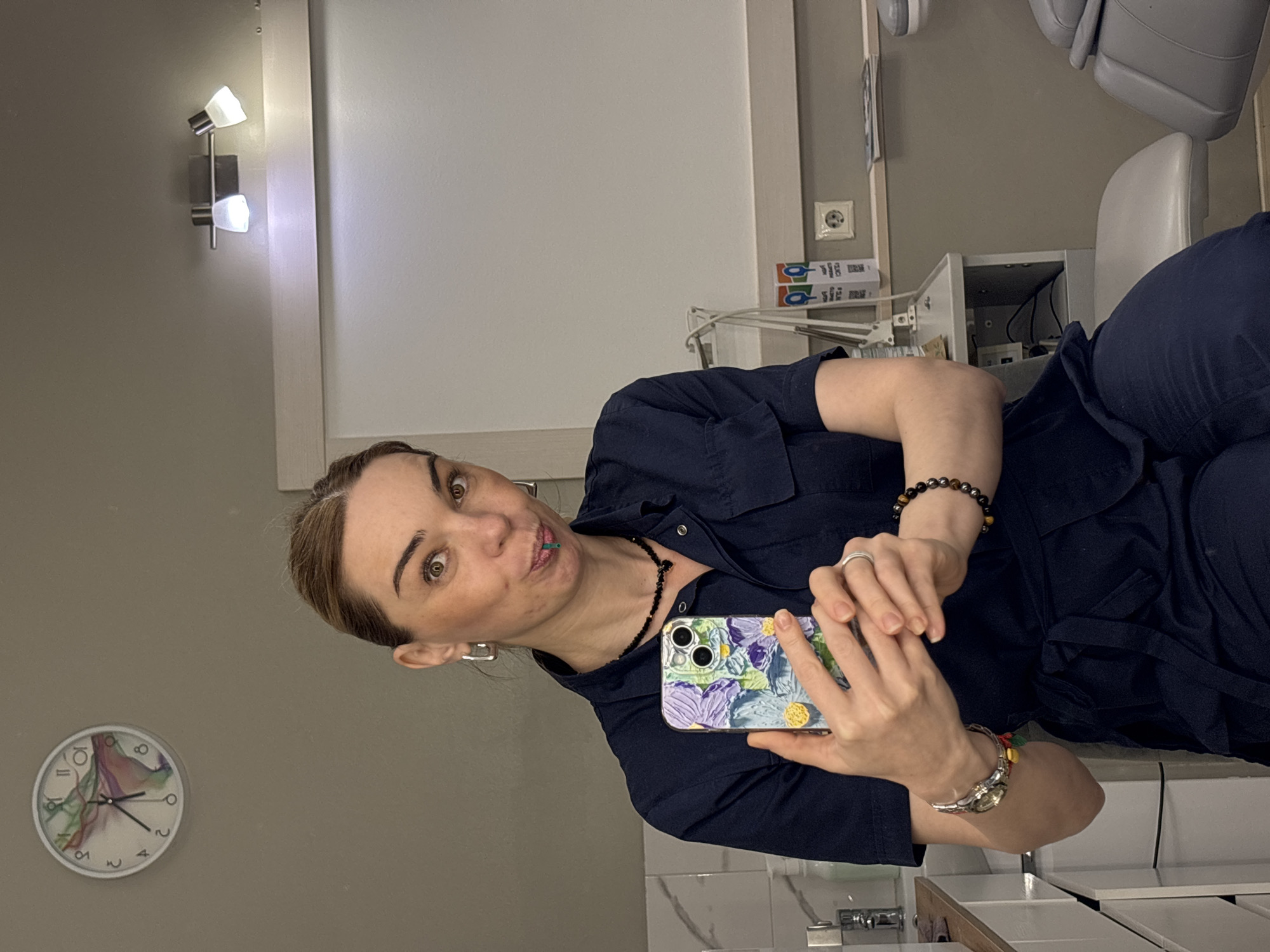 A woman is taking a selfie in a room that appears to be a dentist's office. The time shown on the clock on the wall is approximately 7:06. A dentist chair is located to the right of the frame. The location is likely in the city of Vsevolozhsk, Russia.