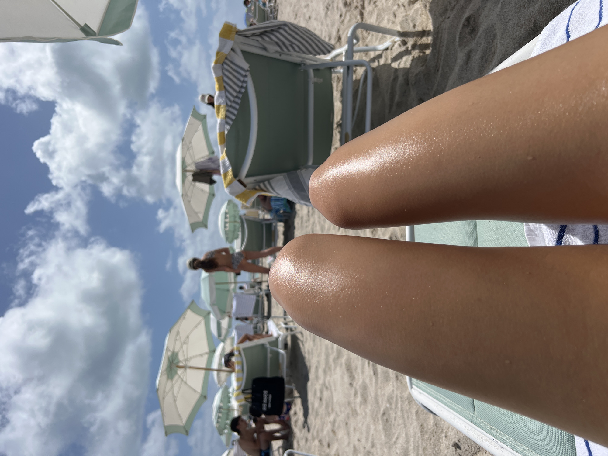 The image shows a beach scene, likely in Miami Beach, United States of America. In the foreground are two sun-tanned legs resting on a beach chair. The skin appears slightly sweaty or oiled. In the background, there are numerous beach umbrellas, people, and chairs scattered on the sandy beach. The sky is blue with puffy white clouds. Some of the beach chairs have striped towels draped over them. People are walking around, some in swimwear. The overall scene suggests a sunny day at a crowded beach.