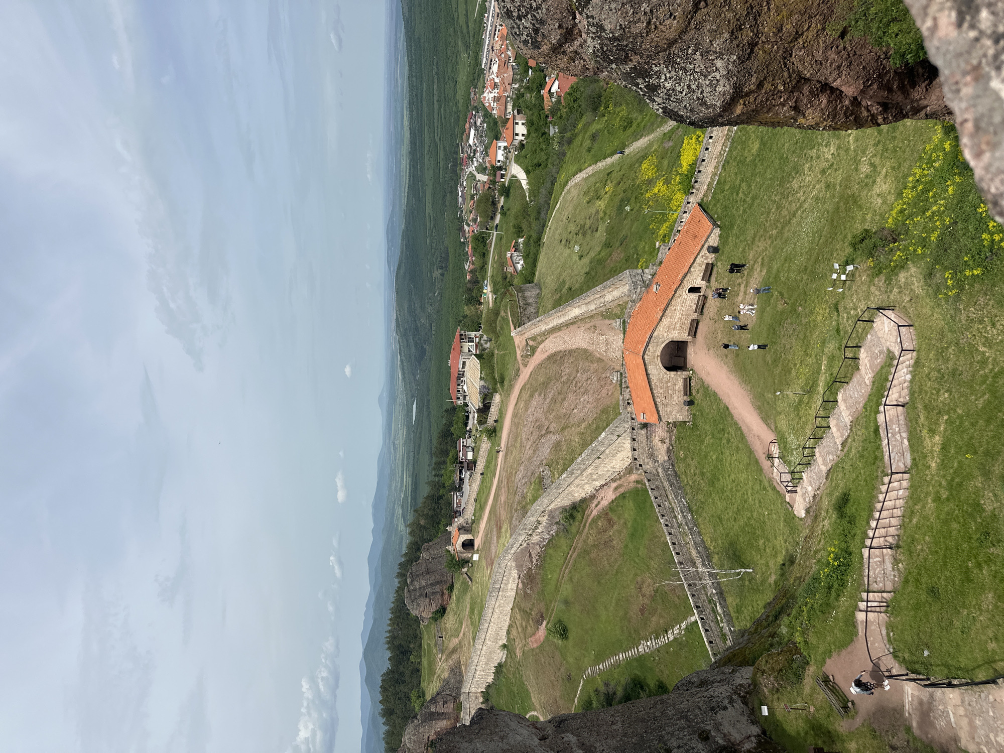 The image depicts a high-angle view of the Belogradchik Fortress in Belogradchik, Bulgaria, a historical structure with stone walls and red-tiled buildings. People are visible within the fortress grounds, some standing or sitting near the buildings. Beyond the fortress lies a town with various buildings, and in the distance, a landscape of mountains and forests stretches out under a cloudy sky. The viewpoint is from a rocky outcrop.