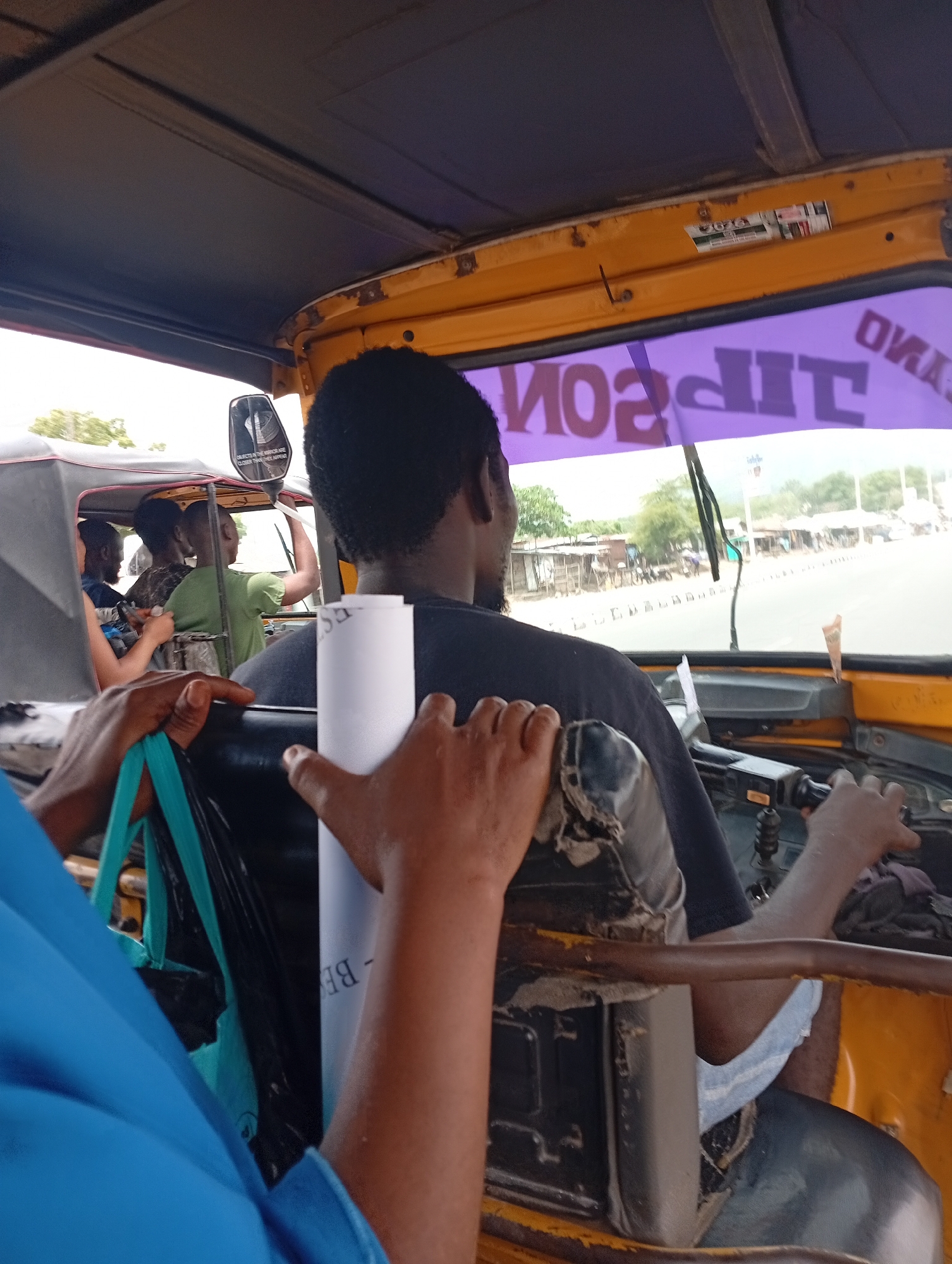 The image depicts the interior of a yellow auto-rickshaw, likely in transit in Adankolo, Nigeria. The driver, a Black man, is visible from the back, wearing a dark T-shirt. His hands are on the steering controls. The windshield has a purple sun visor with text possibly reading "TIPSON," reversed. Visible through the windshield is a street scene, including other vehicles, buildings, and greenery, suggesting an urban or semi-urban environment. Several passengers are visible, some seated in the back of the auto-rickshaw. One passenger in the foreground wears a blue shirt. Their hands are holding a blue shopping bag, and a roll of white paper. The interior of the auto-rickshaw shows wear and tear.