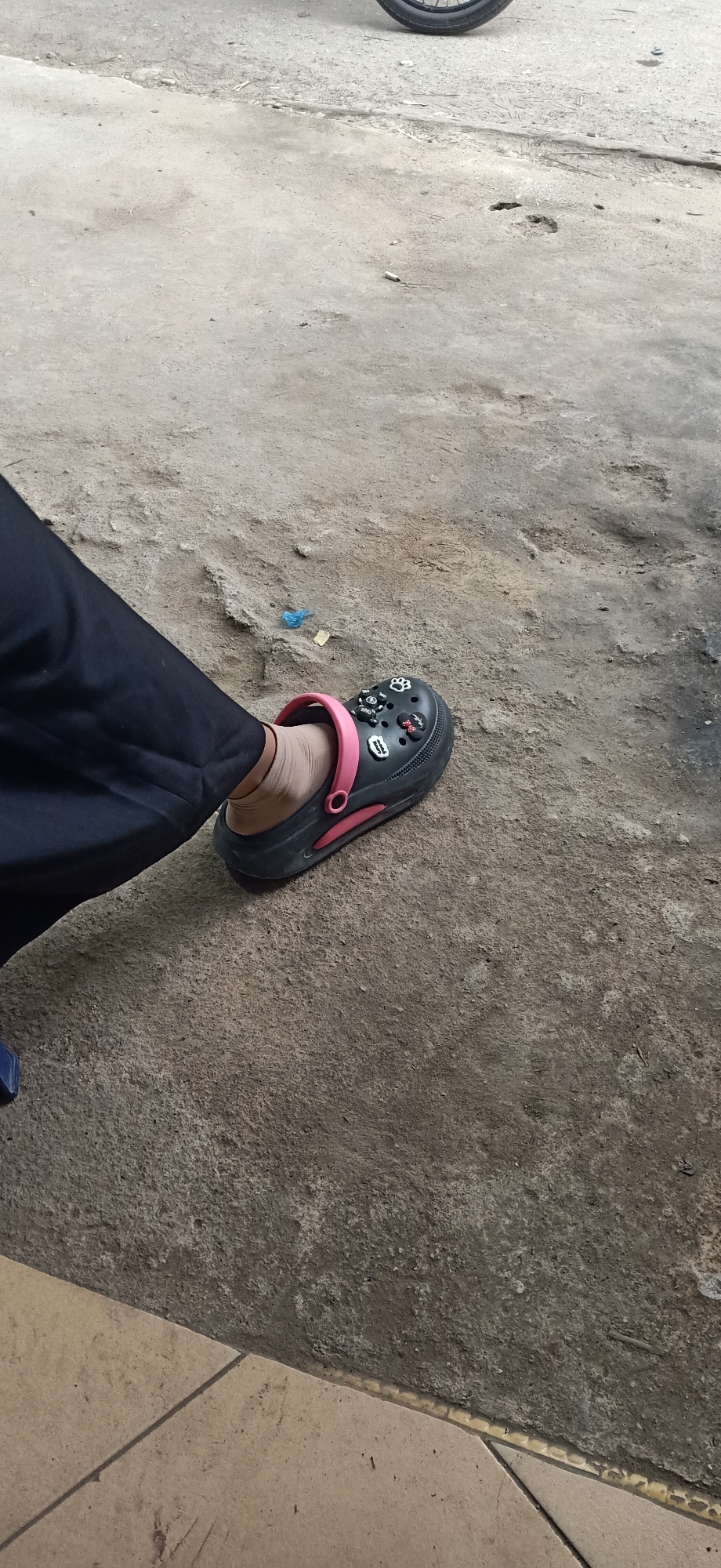 A person's left foot, clad in a black Croc-style sandal with pink accents and various charms, rests on a concrete surface. The sandal is decorated with several small charms, including a paw print, a cartoon character, and what appear to be letters or symbols. The person is wearing a light-colored ankle sock and black pants. The setting appears to be outdoors, with a rough, textured concrete ground. In the background, a portion of a motorcycle wheel is visible. The lighting suggests it is daytime, possibly overcast given the diffused light. The scene is simple and casual, with no strong emotional cues apparent. No discernible text is visible beyond the charms on the shoe.