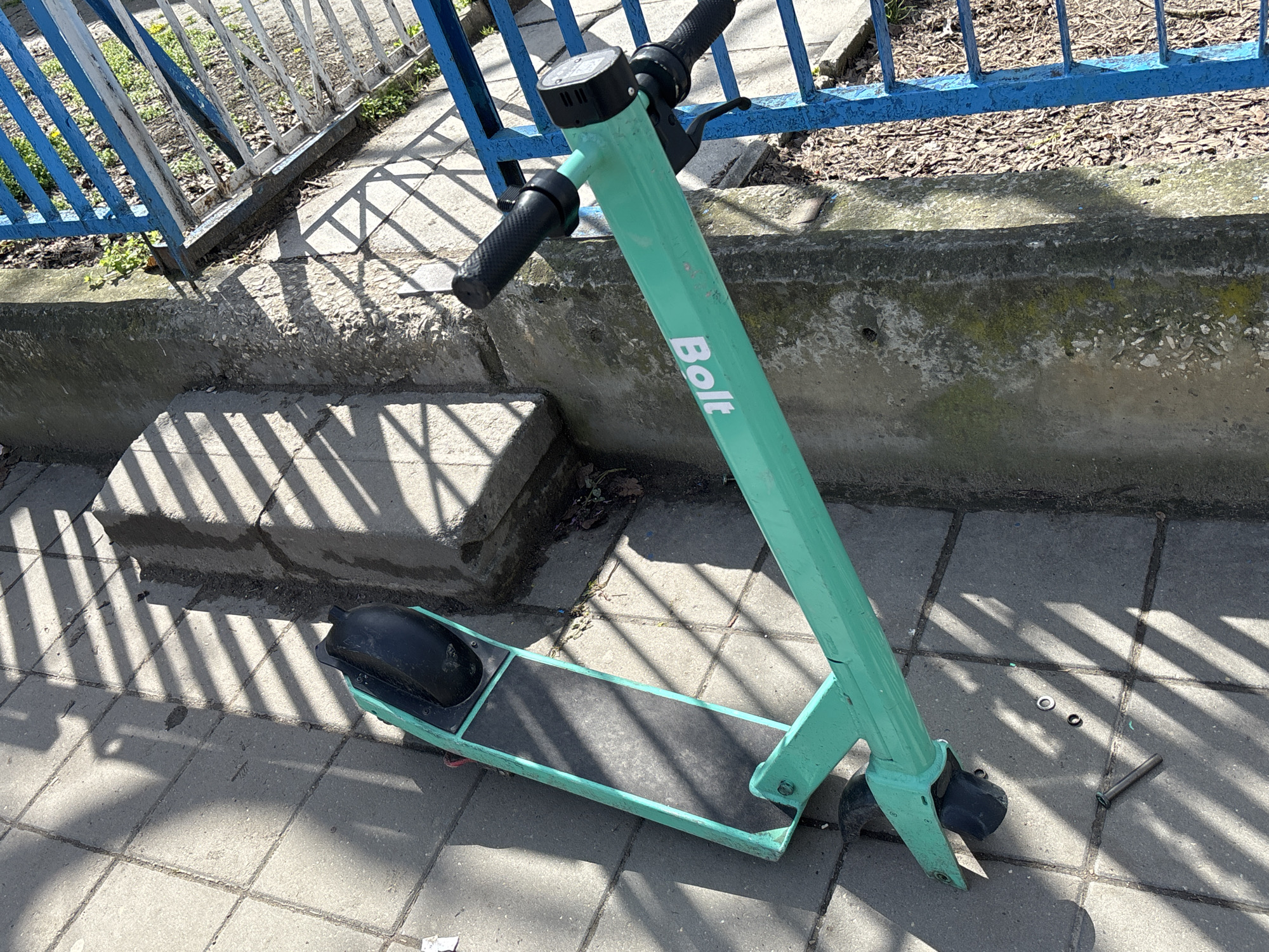 An abandoned Bolt scooter lies on the sidewalk in Varna, Bulgaria. The scooter is light green with "Bolt" branding on the stem. It is missing parts, including the front wheel. The scooter is leaning against a concrete curb and a blue and white metal fence. The sidewalk is made of gray paving stones and is partially covered in shadows from the fence. Debris, including metal components from the scooter, are scattered near the front of the scooter.