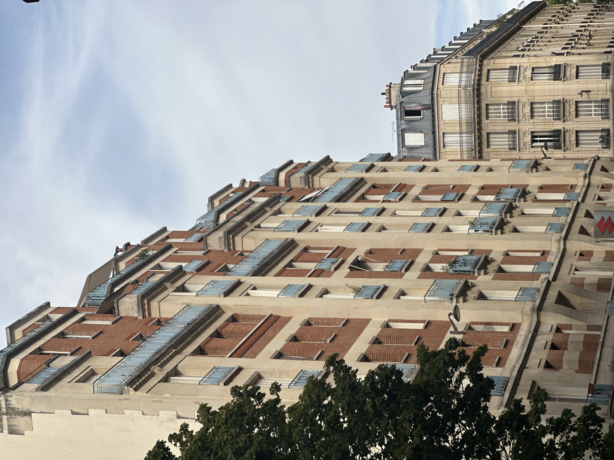 This is a medium-shot, eye-level, vertical-format photograph of two apartment buildings in Paris, France, taken on a cloudy day. The building in the foreground is to the left, taller, and features a facade of light beige stone with brick accents and light blue balconies. The building in the background to the right is slightly shorter and features a mansard roof with dormer windows, also made of light beige stone. The buildings are typical of Parisian architecture, with their multi-story facades, numerous windows, and ornate detailing. The scene is devoid of people and any visible actions or interactions. The environment is an urban street, with a large green tree on the left side of the frame obscuring part of the foreground building. The sky is overcast with some wispy clouds.
