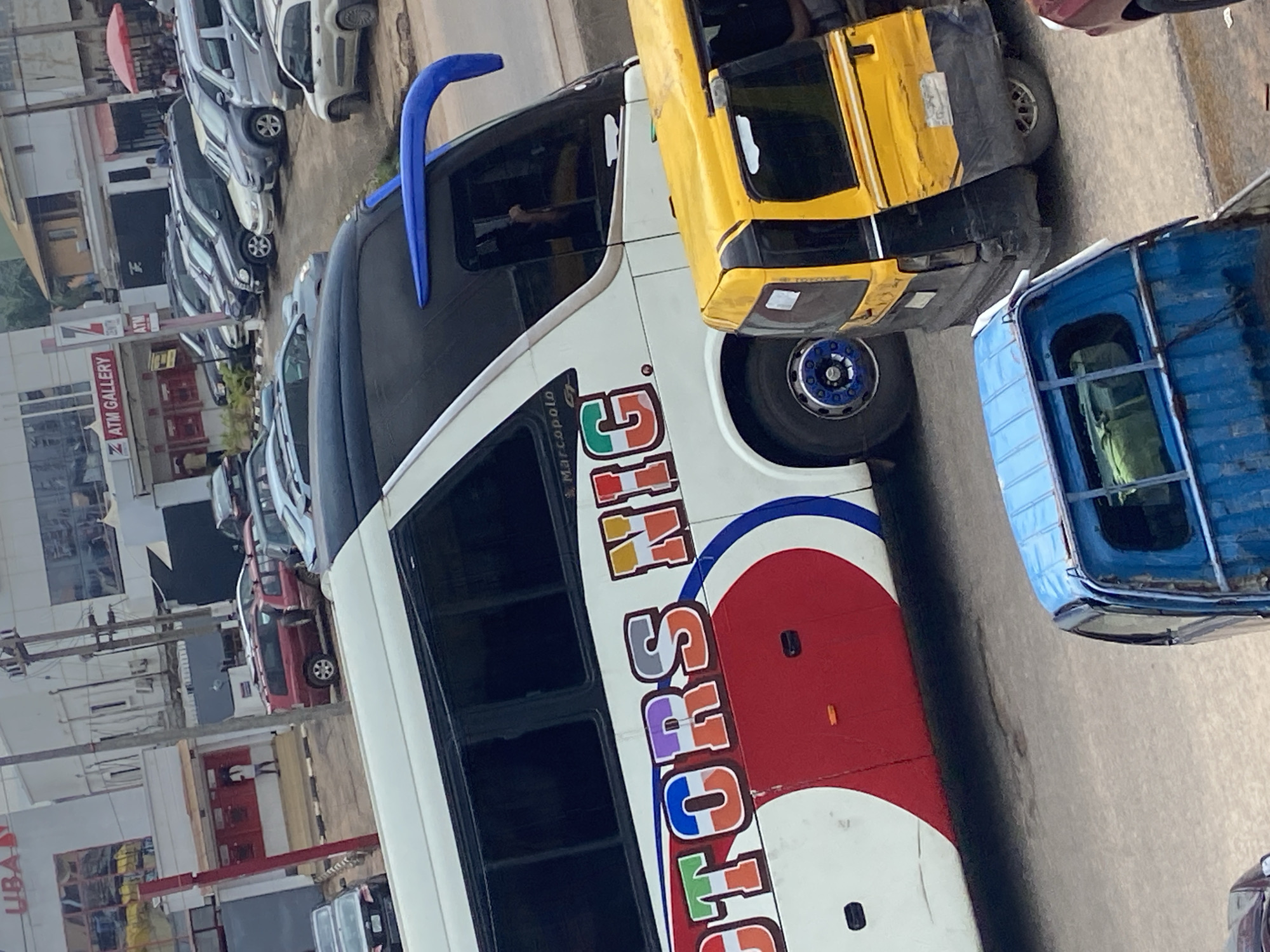 The image depicts a street scene in Ogbunike, Nigeria, with a variety of vehicles and buildings. In the foreground, a large white bus with "MOTORS NIG" emblazoned on its side is prominently featured. The bus has a red and white design on its lower half and a black roof with a blue aerodynamic spoiler. To the right of the bus, a yellow auto-rickshaw with its door open is parked. Further in the background, several other vehicles, including SUVs and cars, are visible, suggesting a busy commercial area. Buildings with commercial signage, such as "UBA" and "ATM GALLERY," line the street, indicating a financial or retail district. A person is seen standing near the red entrance of a building, and other individuals are present in the background, suggesting daily activity in this urban environment. The overall scene captures a typical street view in a Nigerian city.
