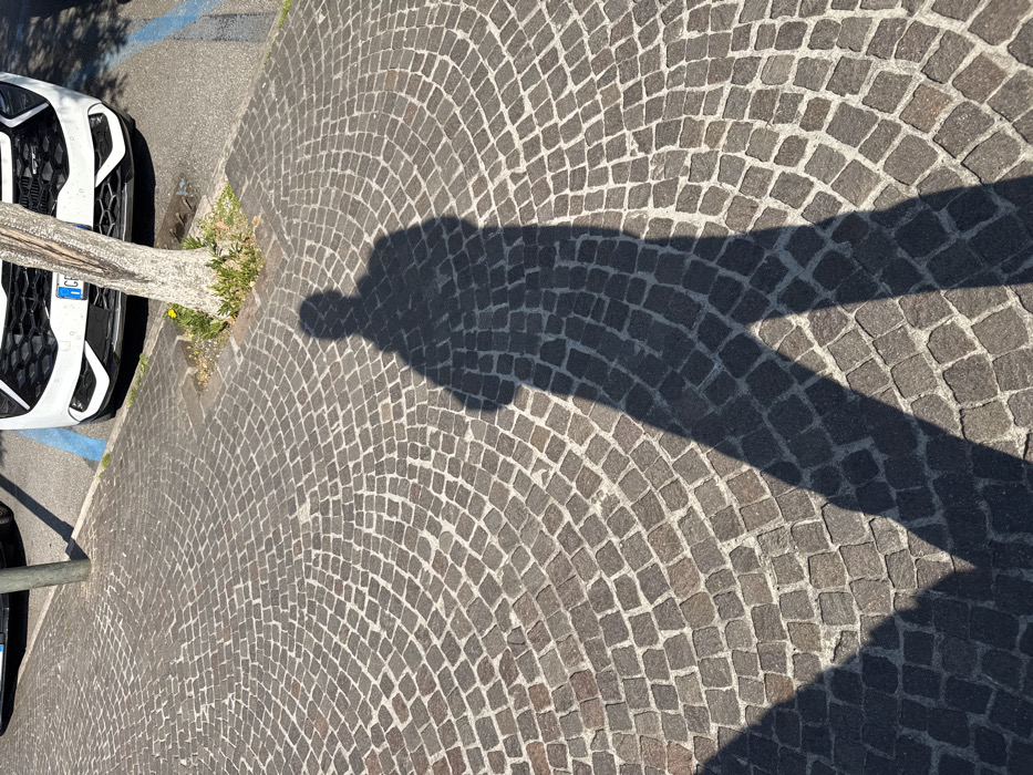 The scene depicts an outdoor urban environment in Sirmione, Italy, under bright, direct sunlight. The foreground and midground feature a paved surface composed of dark grey and brown cobblestones laid in a radiating, fan-like pattern. A prominent, dark human-shaped shadow extends from the upper-left quadrant towards the bottom-right of the frame, indicating the presence of an individual standing or walking out of frame to the left. In the background, a white vehicle, specifically its front bumper and grille, is parked on dark asphalt. An Italian license plate, partially visible and beginning with "CF", is affixed to the vehicle. A blue painted line on the asphalt indicates a parking space. A light-colored tree trunk grows from a small patch of earth containing sparse green vegetation, situated between the cobblestone pavement and the parked vehicle. A dark, slender pole is partially visible behind the tree and to the left of the white car.