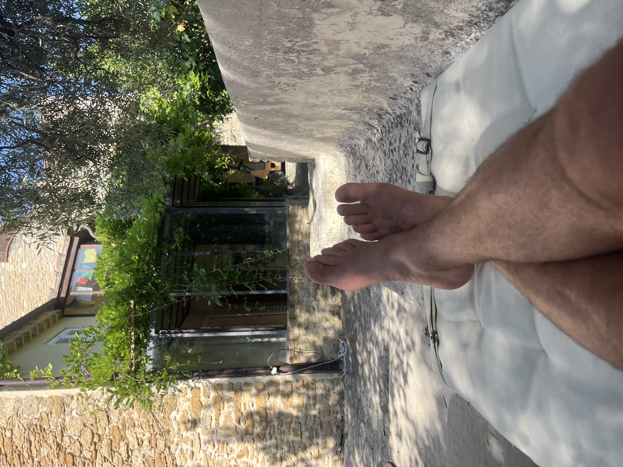 The image depicts a personal perspective of someone relaxing outdoors in Le Castellet, France. The foreground is dominated by the lower legs and feet of an individual, who is lying on a light-colored padded surface. Their legs are crossed at the ankles, and their feet are bare.
The background reveals a rustic setting with a stone building constructed from rough-hewn, light brown stones. A large glass-paned door or window is visible on the building, partially obscured by abundant green foliage and climbing vines with small purple flowers. Above the windows, a section of the building's roof with terracotta tiles is visible, along with a blue sky.
To the right of the building, a weathered, textured concrete wall curves along the scene, creating a natural seating or lounging area. The ground in front of the building and extending towards the concrete wall is paved with irregular stone or concrete slabs, dappled with sunlight and shadow. Mature trees with silvery-green leaves, likely olive trees, cast shadows over parts of the scene, contributing to the warm, sunlit atmosphere. The overall impression is one of peaceful, leisurely repose in a Mediterranean environment.