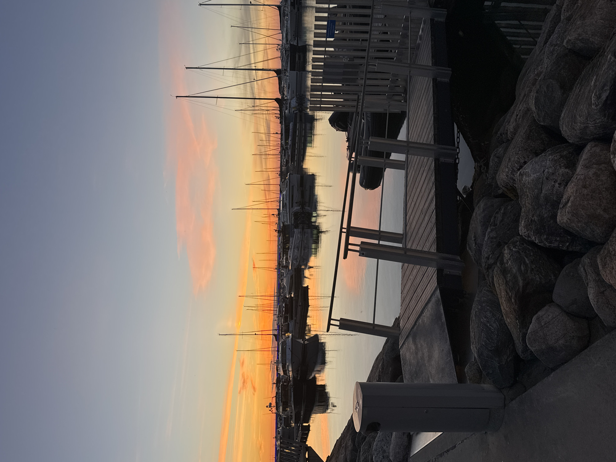 The image depicts a tranquil marina scene at sunset in Tallinn, Estonia. The sky transitions from a soft blue to warm hues of orange and pink, reflecting beautifully on the calm water. A row of sailboats and yachts are moored side-by-side, their masts creating a striking silhouette against the colorful sky. The wooden dock is visible in the foreground, with a railing and a waste receptacle. Large, weathered rocks are piled along the edge of the marina, adding a natural element to the man-made structure. The overall atmosphere is serene and peaceful, suggesting the end of a pleasant day. The time of day is clearly sunset. There is no visible text in the image.
