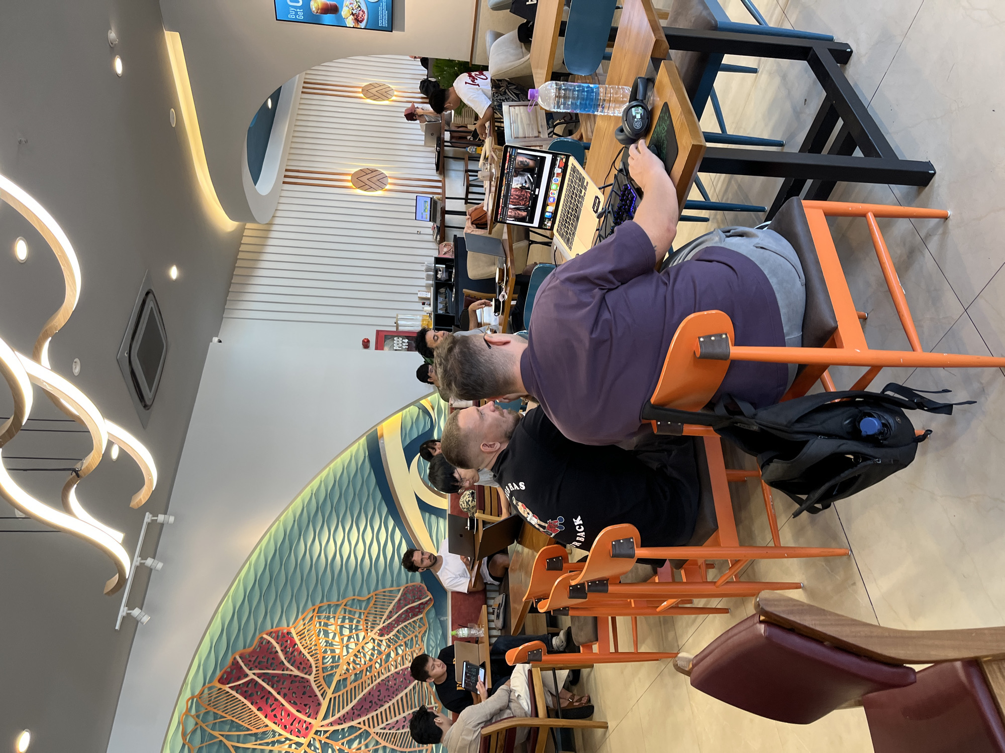 The image captures the interior of a cafe or co-working space bustling with activity, likely in Đà Nẵng, Vietnam. People are seated at shared tables, predominantly working on laptops and tablets. A man in a purple shirt and grey pants is at a table with a laptop; another man in a black shirt with a basketball graphic sits nearby. There are multiple people working at the long table in the background; one person is wearing a white shirt. Backpacks are visible on the backs of chairs and on the floor. Tables are made of wood, with a dark metal frame on some. Chairs are predominantly orange. A water bottle and headphones sit atop a laptop. The lighting is bright. A large decorative wall art piece featuring a stylized leaf design in teal and orange can be seen on the left wall. Ornate light fixtures hang from the ceiling. A flat screen displaying an advertisement is positioned higher up on the wall. The floor is tiled in a light color. The scene suggests a casual and productive atmosphere.