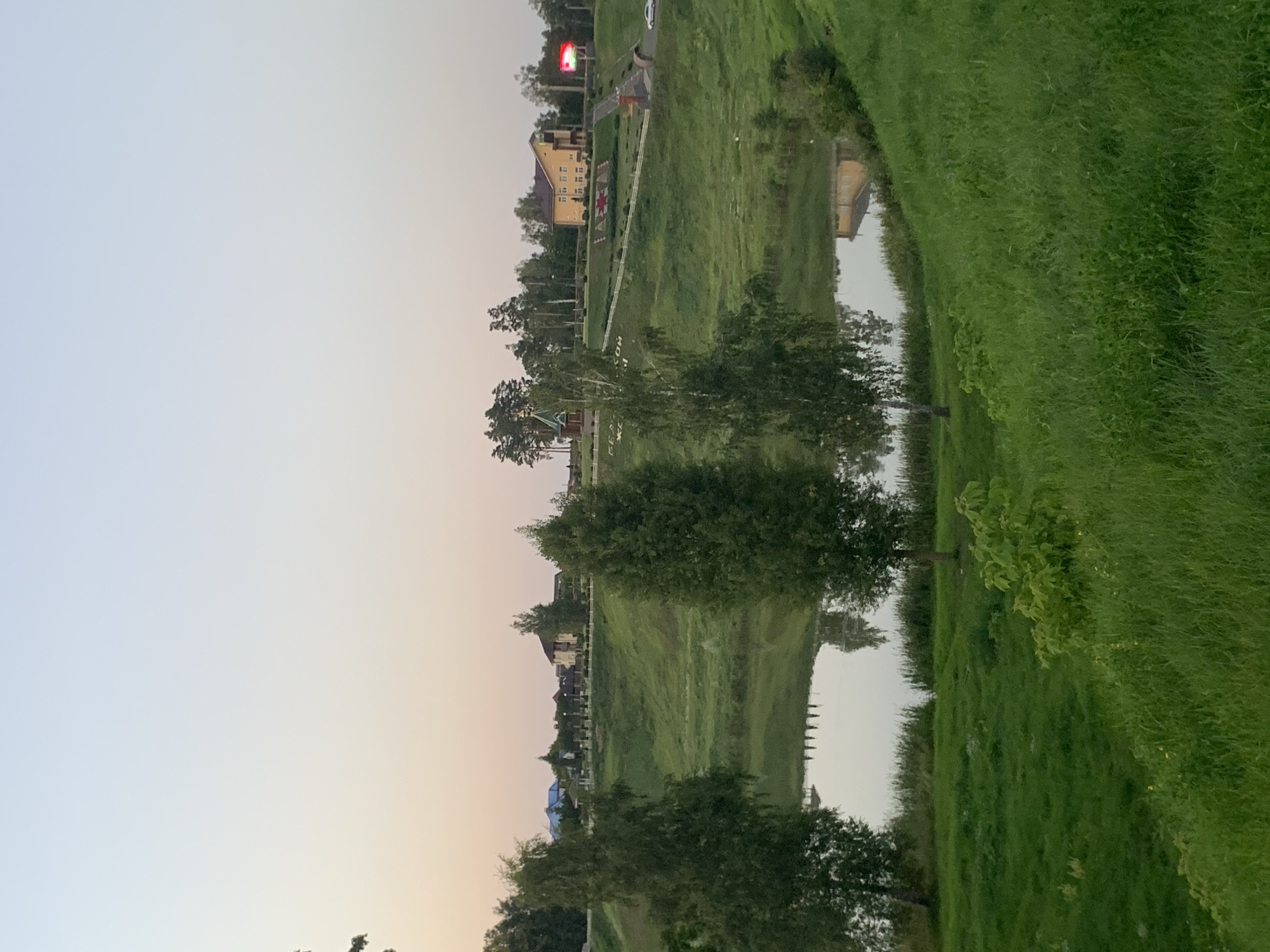 The image depicts a serene landscape in Kapyĺ, Belarus, featuring a body of water reflecting the pale sky of dusk. The foreground is dominated by lush green grass covering rolling hills. In the mid-ground, a calm lake or pond is visible, its surface mirroring the trees on its banks. Several trees, including two prominent birch trees with distinctive white bark, stand on the edge of the water. Further in the background, a green, grassy embankment rises, dotted with more trees and several residential buildings. One building, a light yellow, two-story structure with a red roof, stands out, as does a smaller red-roofed building resembling a chapel with a green dome. A red LED billboard is also visible on the right side of the background. A white car is parked on a road in the distance, and a white fence lines a path on the embankment. The sky transitions from a pale blue at the top to a soft pinkish hue near the horizon, indicating either sunrise or sunset.